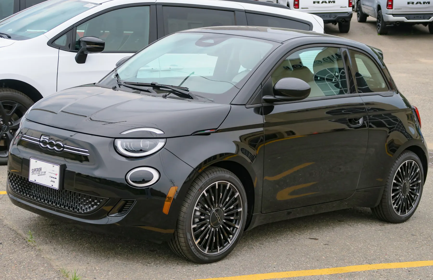 Front three-quarter of a glossy black Fiat 500e under an overcast sky, with a chrome 500 badge centered in thin horizontal chrome bars across the black nose, round headlamps with bright LED rings and a separate thin chrome eyelid arc on the hood edge above each, a second round ring-lit lamp in the bumper, a separate amber side marker at the bumper corner, a black mirror cap, a black window surround, a body-colored flush door handle, a machined silver wheel with black pockets, a dealer placard reading Superior Chrysler Dodge Jeep Ram, and a white vehicle and Ram pickups behind it