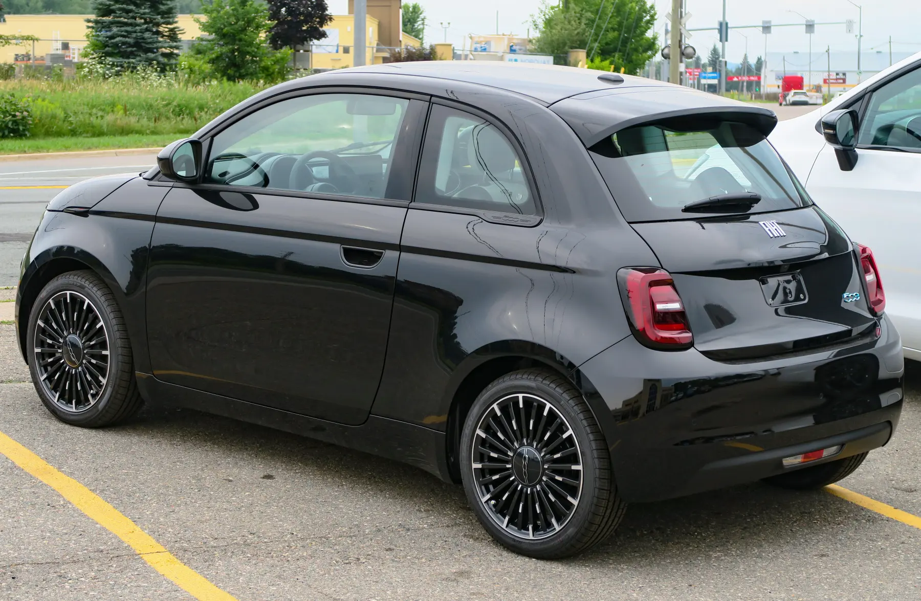 Rear three-quarter of a glossy black Fiat 500e, with chrome FIAT lettering above an empty plate recess that holds a bracket, a small blue 500e badge to the right of the recess, a red taillamp with a clear white bar inside, a wiper on the rear window, a machined silver wheel with black pockets, and trees, a yellowish building, and street poles and signs behind it