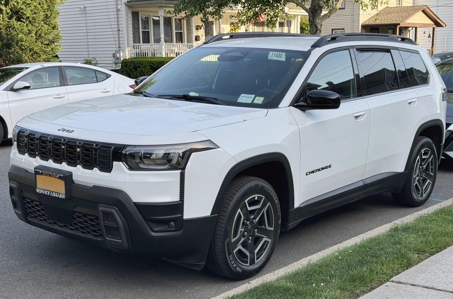 Front three-quarter of a white Jeep Cherokee on a residential street, nose to the left. A Jeep script sits on the hood above seven dark grille slots. The headlamp is a wide squared housing with an angular element in the clear lens and an amber strip at the outer upper corner. A black ribbed strip runs down the body just outside the lamp. The lower bumper and the wheel-arch cladding are black. Black capital letters spelling CHEROKEE are on the front door. Black roof rails run above the glass. The plate frame reads NEW YORK and SERVICE LOANER. A window sticker is on the windshield. A pale house with a porch and an American flag is behind, and a white sedan is parked at the left. Grass and a sidewalk are in the foreground.