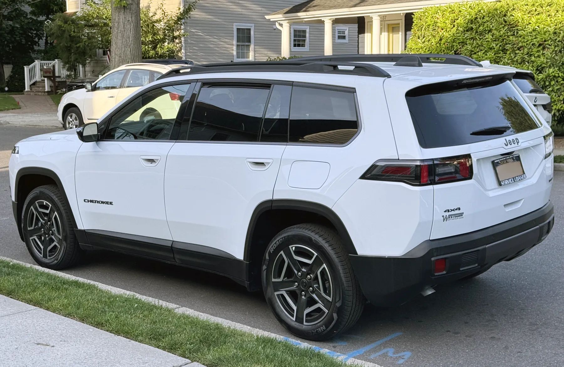 Rear three-quarter of a white Jeep Cherokee on a residential street, tail to the right and flank to the left. Black capital letters spelling CHEROKEE sit on the front door. Black roof rails run above the side glass. The wheel arches and the rocker are black, and the arches are squared. A dark horizontal tail lamp crosses the seam between the quarter panel and the hatch, with red segments in the lens. The word Jeep is centered on the hatch above the plate. The hatch reads 4x4 on the lower left, with a small dealer decal beneath it. The plate frame reads NEW YORK along the top and SERVICE LOANER along the bottom. The wheels are dark with bright machined edges. A pale house with a columned porch, a tree, a hedge, and a white SUV are behind. Grass and a sidewalk are in the foreground.