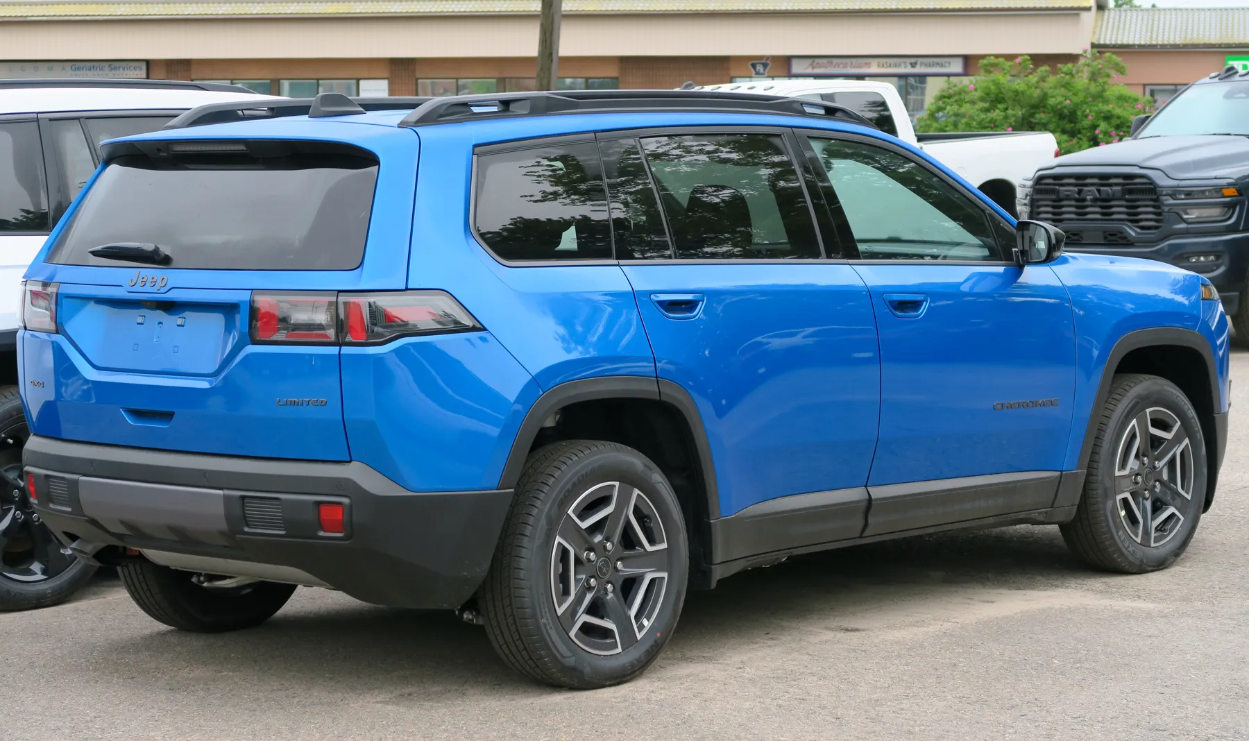 Rear three-quarter of a bright blue Jeep Cherokee in a dealer lot, tail toward the left and flank running to the right. The tail lamp is a dark horizontal housing split by the hatch edge, with red segments and a clear section in the lens. The word Jeep is centered on the hatch. Spaced capital letters spelling LIMITED sit on the rear quarter below the lamp. CHEROKEE is spelled in dark letters on the front door. Black roof rails, black squared wheel arches, and a black lower bumper with a small red reflector. A white SUV and a dark Ram pickup are parked nearby. A low commercial building with storefront signs is behind.