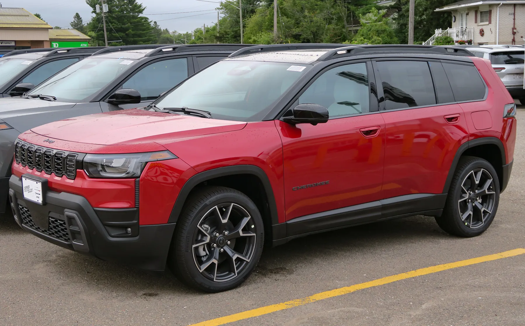 Front three-quarter of a red Jeep Cherokee in a lot, nose to the left, with water spots on the paint. A Jeep script sits on the hood above seven dark grille slots. The headlamp is a wide squared housing with an amber segment at the outer corner, and a black ribbed strip runs down beside it. CHEROKEE is spelled in dark letters on the front door. The wheel arch is a squared black cap, and the lower bumper and rocker are black. Black roof rails run above the glass. The wheels are dark with machined edges. A dealer plate is on the front bumper. Gray and silver SUVs are parked alongside, a yellow line crosses the asphalt, and trees, a house, and a low commercial building are behind.