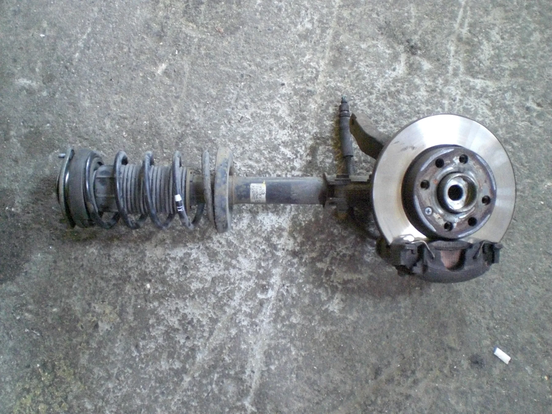 Coil spring wrapped around a damper tube on a suspension unit, knuckle, brake disc, and caliper still attached, sitting on concrete
