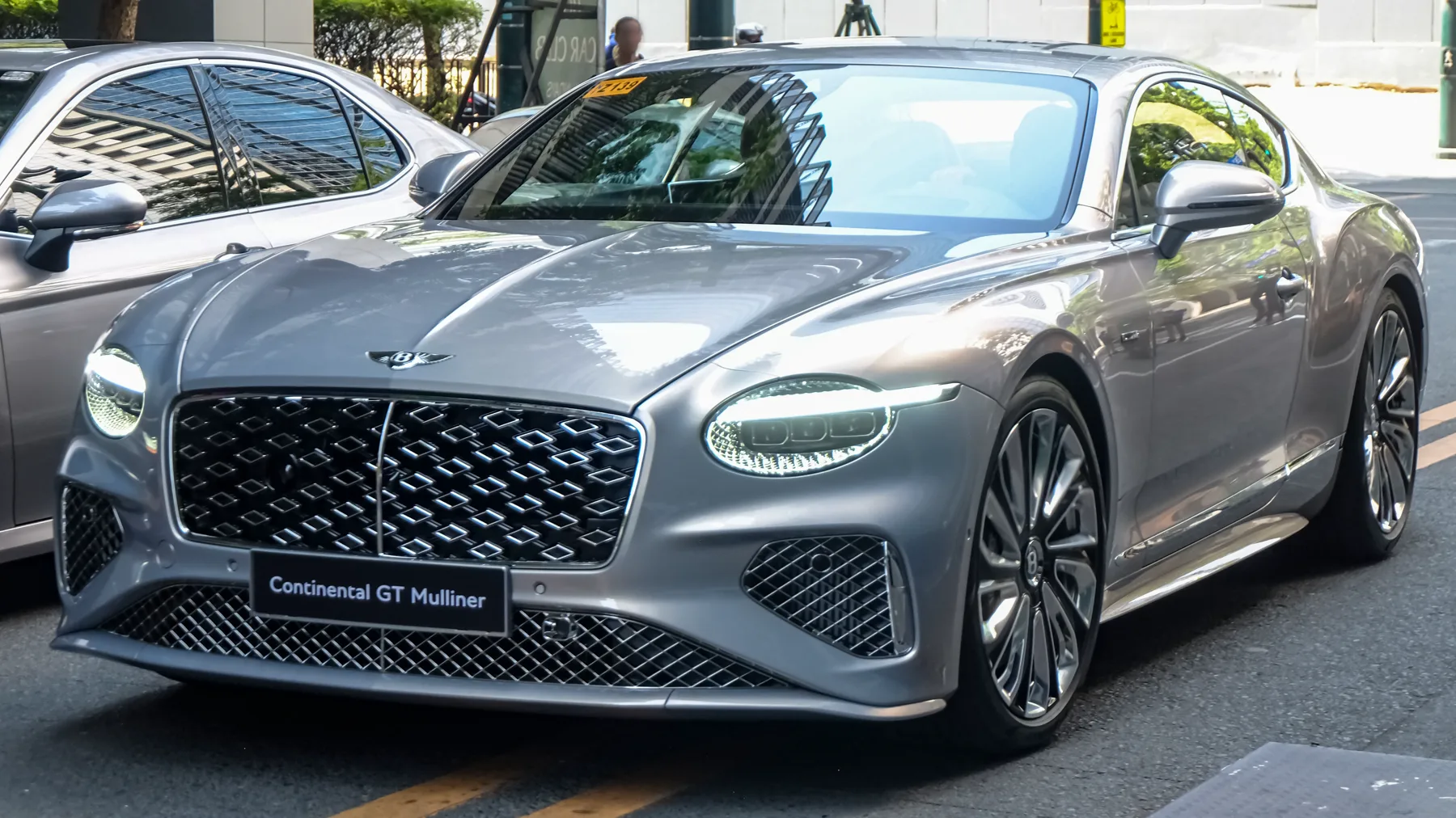 Front view of a metallic silver-gray Bentley Continental GT outdoors on a city street, asphalt with a yellow road line. The nose points toward the lower left and the flank runs back to the right. Another gray car is partly visible at the left edge. Both headlamps are visible, each a single oval with an LED strip along the upper edge. The grille is a bright chrome floating-diamond mesh with a chrome centre bar. A black plate on the bumper reads Continental GT Mulliner. The mirrors are body-color silver-gray. The wheels are two-tone dark-and-silver multi-spoke.