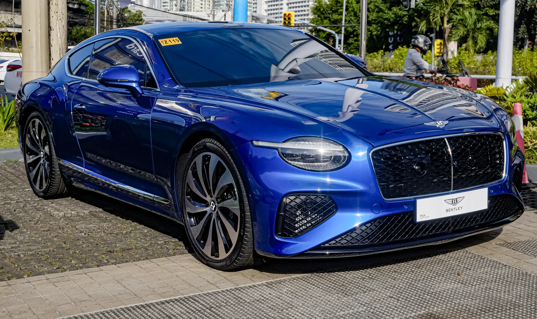 Front three-quarter of a vivid deep metallic blue Bentley Continental GT outdoors on a paved lot, nose to the right of the frame, flank running back to the left. Trees, a traffic light, a building, and a motorcyclist are in the background. A white car is partly visible at the far left edge. A chrome winged B sits on the hood. The headlamp is one oval with an LED strip along its upper edge. The grille is a black diamond-pattern mesh with a thin chrome vertical centre bar and a chrome surround. A white dealer plate on the bumper reads BENTLEY. The lower intakes are black diamond mesh. The mirror is body-color blue. The wheels are two-tone dark-and-silver multi-spoke.