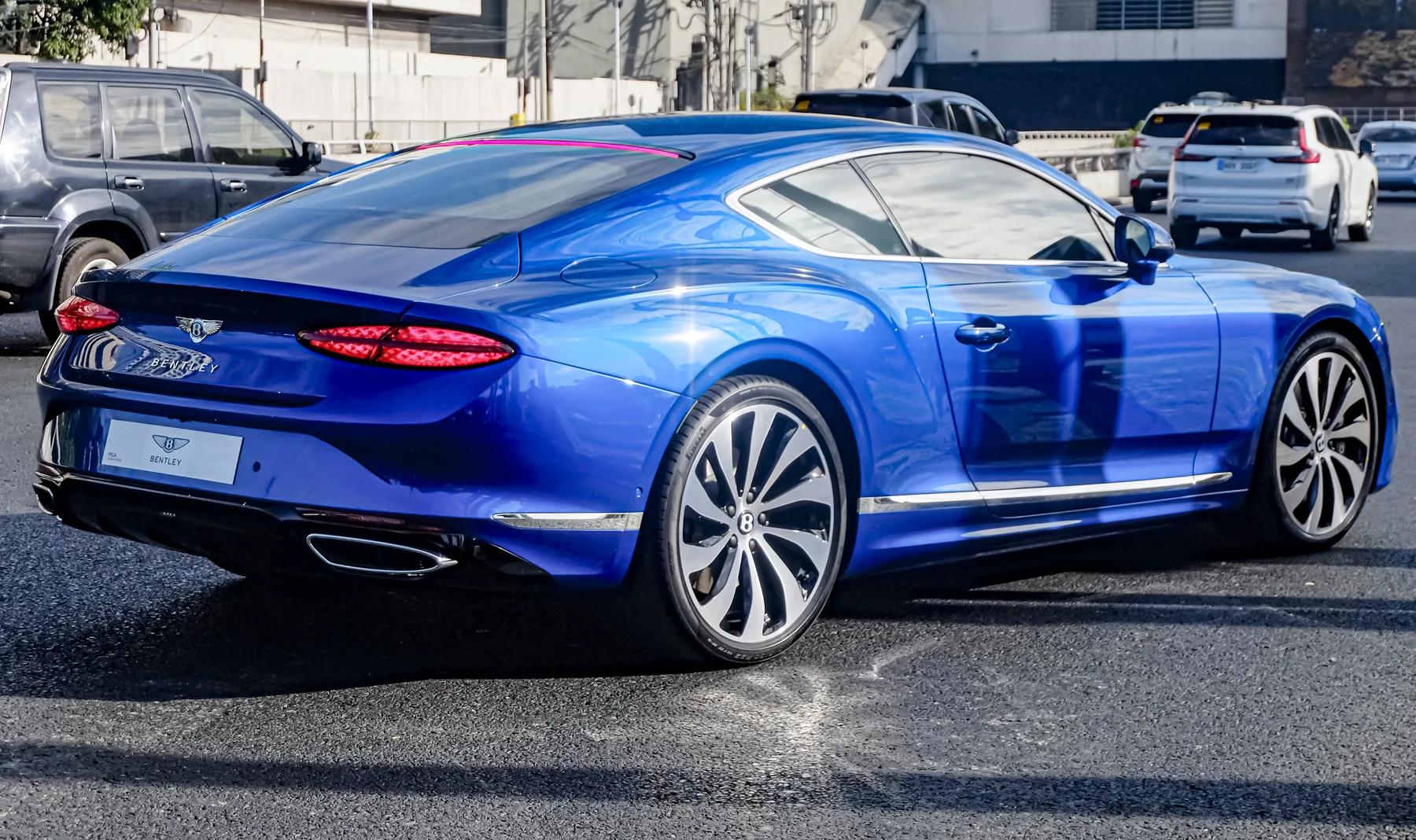 Rear three-quarter of a vivid deep metallic blue Bentley Continental GT outdoors on an asphalt road. A dark SUV is at the left, and white SUVs and cars sit behind at the right. The trunk is at the left of the frame; the flank and wheels run to the right, with the rear wheel to the right of the trunk. Chrome letters spelling BENTLEY and a winged B badge are on the trunk. The taillamps are slim, red, horizontal slashed-oval clusters. One oval chrome exhaust outlet is clearly visible at the lower left of the bumper. A white dealer plate on the bumper reads BENTLEY. The wheels are two-tone dark-and-silver multi-spoke.