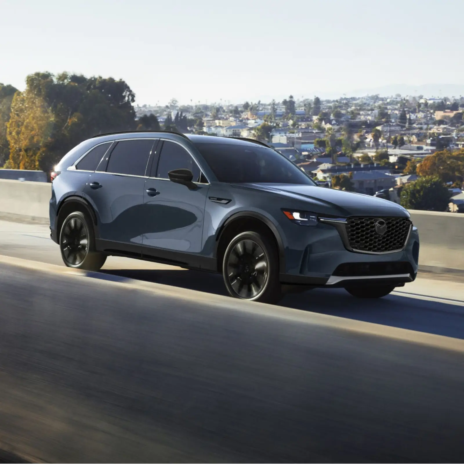 Dark 2026 Mazda CX-90, front three-quarter on a highway with the Signature Wing spanning the grille into the slim lamps