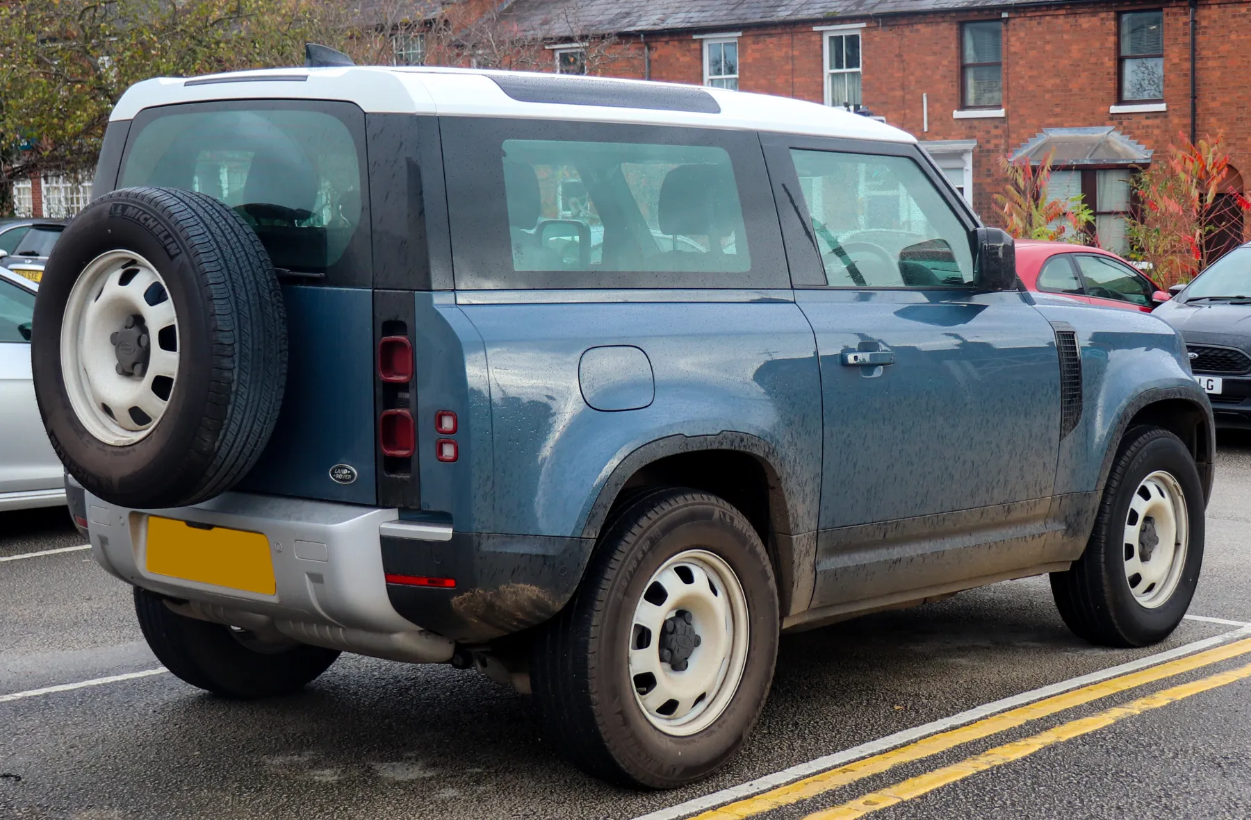 Rear three-quarter of a blue 2022 Land Rover Defender 90 with a white roof, steel wheels, a spare wheel on the side-hinged rear door, and a vertical tail lamp at the rear corner