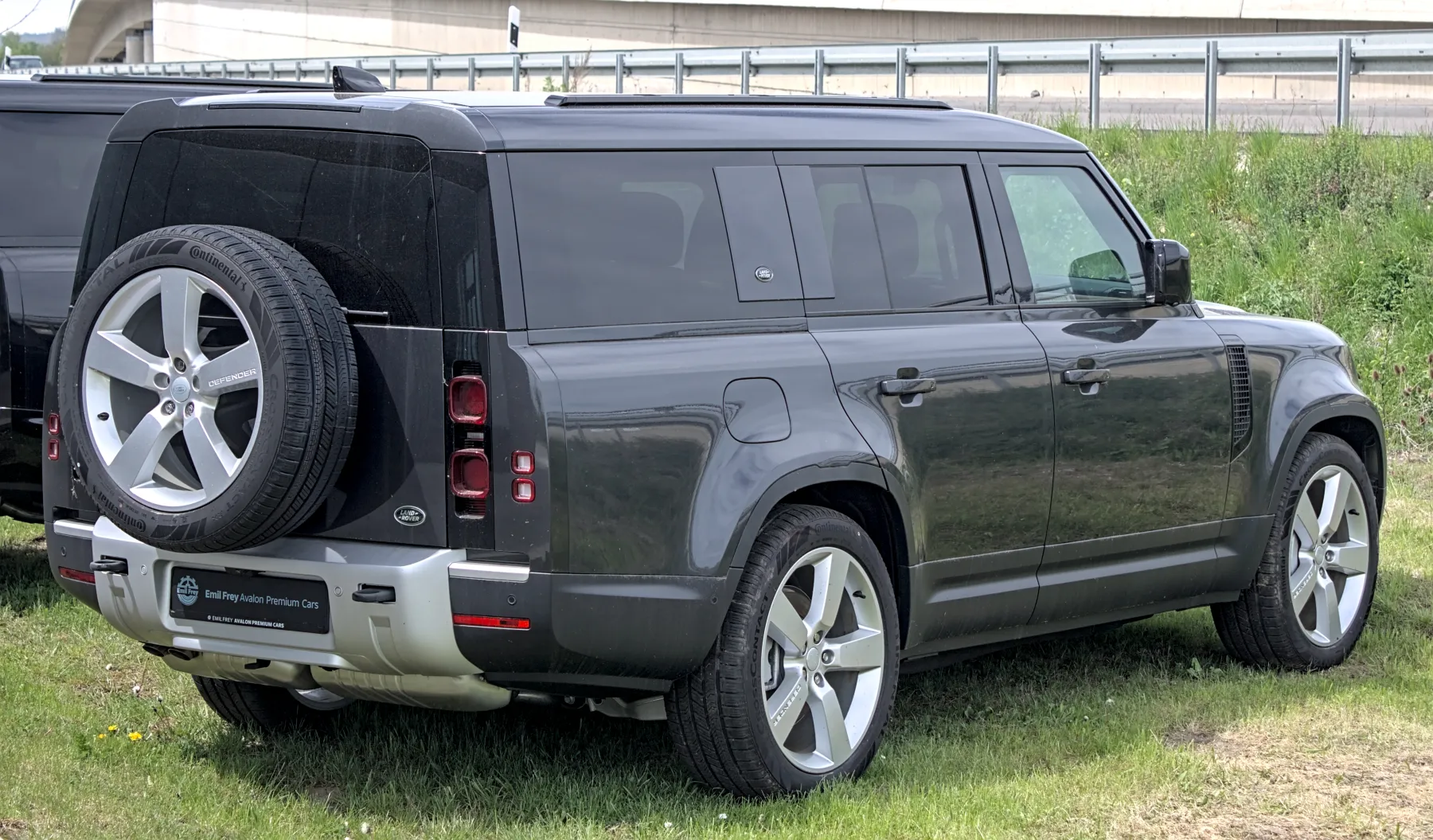 Rear three-quarter of a dark grey Land Rover Defender 130 with a black roof, an alloy spare on the rear door, vertical tail lamps, and a long side with an extra rear quarter window