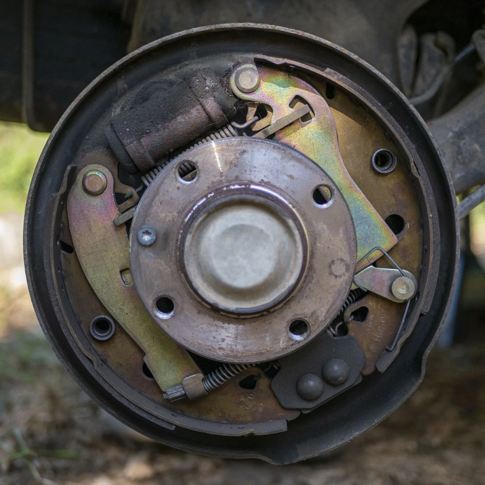 Outdoors, still on the car. The background is blurred dirt ground and greenery. The car's rear axle arm is at the upper right. In the center is a hub flange with a domed center cap and four empty bolt holes, and no wheel studs are visible. The dark, rusted wheel cylinder sits at the upper left, with a coil spring running right just under it. The two shoe webs are gold-tinted zinc-plated steel, and the linings are hidden behind the webs at the rim. At the upper right is a gold-tinted adjuster lever and quadrant on a pivot pin. At the right is a lever end with a pin and a thin wire clip. At the bottom are a lower coil spring and a dark gray anchor block with two round bolt heads.
