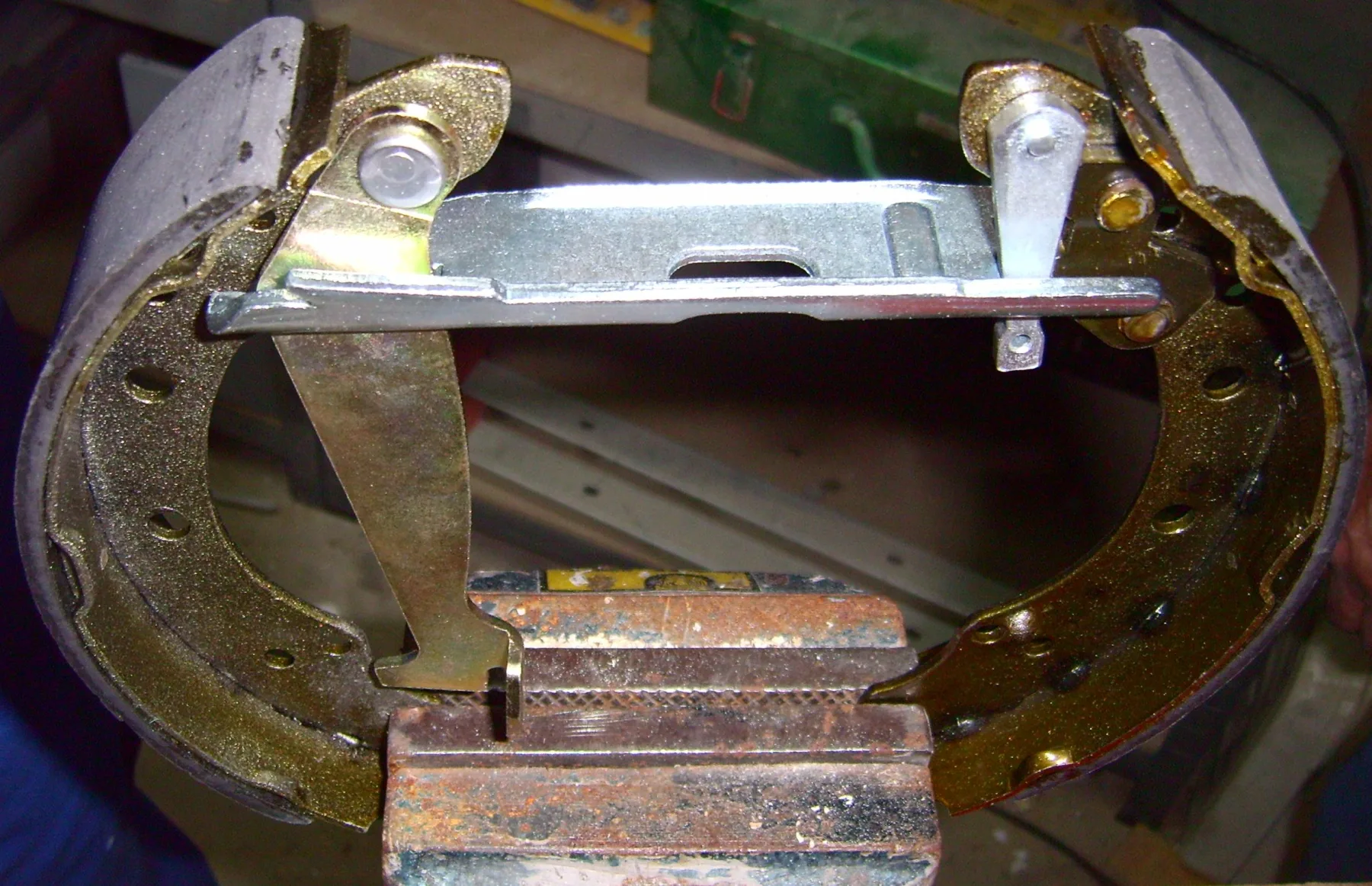 The two shoes are clamped upright in a rusty bench vise in a workshop, with a green metal toolbox behind. The shoe webs are gold-tinted with drilled holes, and the gray linings are on the outside. A flat silver strut bar with a slot spans the top between the shoes. A gold parking lever hangs down from a pivot pin on the left shoe, with a hooked lower end. A small silver piece hangs down near the right end of the strut. No cable and no coil spring are visible, and rivets cannot be confirmed.