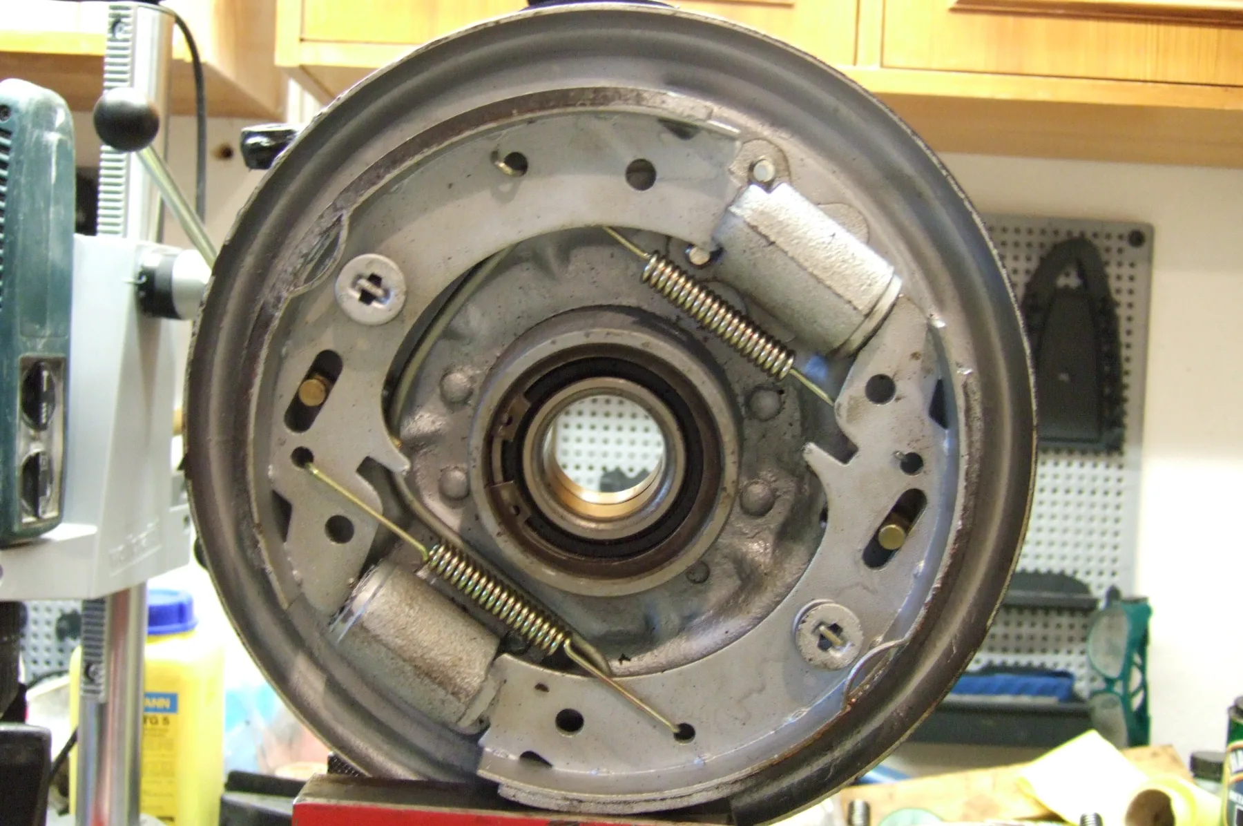Off the car, standing upright in a home workshop on a red-painted stand. A drill press is at the left, with a pegboard wall and light wooden cabinets behind. The backing plate is light gray and silver. Two silver-gray wheel cylinders sit diagonally opposite each other, one at the upper right and one at the lower left. Each has a coil spring and a wire rod running diagonally from it. No rubber boots and no brake lines are visible. The center is a hub opening with a bearing and seal, and no wheel studs are visible. Small screw heads hold the shoes at the upper left and lower right.