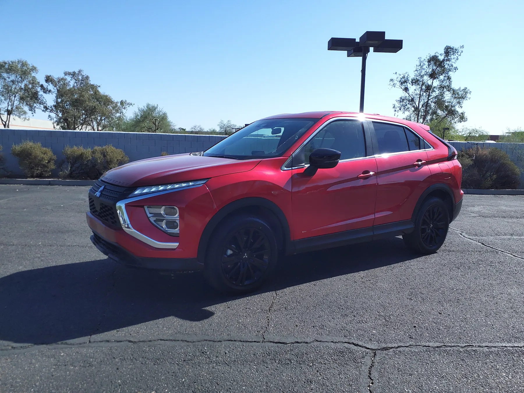 Front three-quarter of a red 2023 Mitsubishi Eclipse Cross LE with black wheels, alone on an empty lot with a block wall and shrubs behind, the three-diamond emblem in the grille, and lettering along the leading edge of the hood
