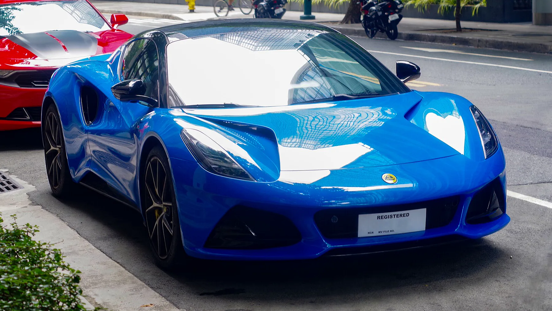 Front three-quarter of a bright blue Lotus Emira from the front right, nose to the right, parked at a curb on a city street. The headlamps are dark. The hood duct above the headlamp nearest the camera is a dark recess; the other duct is lost in sky reflection on the hood. A round yellow-and-green Lotus badge sits on the blue nose above the plate and the lower intake. The lower nose is a black mesh intake, and a white plate reads REGISTERED. The wheels are black multi-spoke with yellow calipers. A large black intake is behind the door. The roof and mirror are black. A red car with a black stripe on the hood is at the upper left, with motorcycles and a bicycle along the sidewalk.