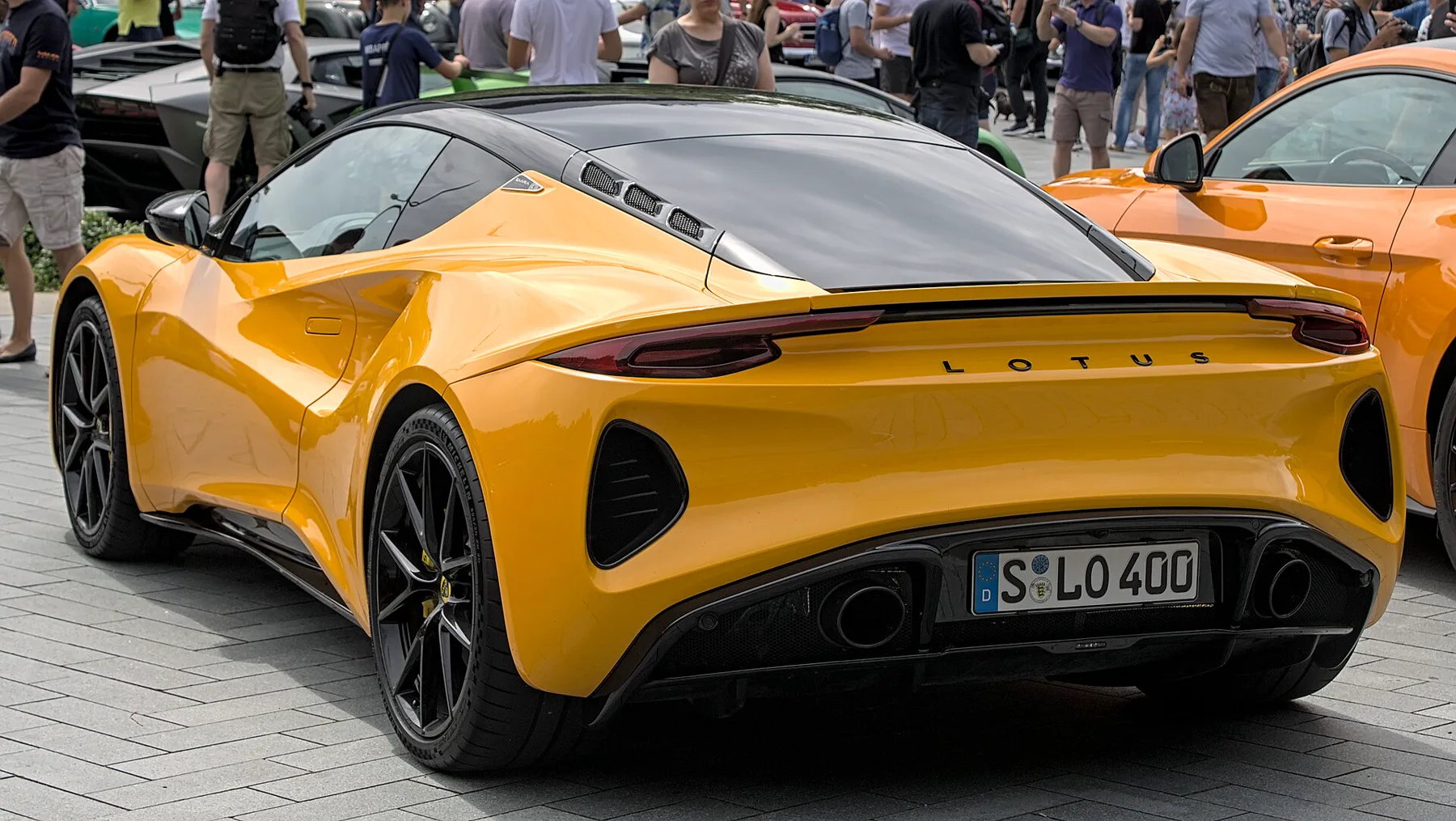 Left rear of a golden yellow Lotus Emira on gray block paving at a crowded car meet, with many people standing behind it. An orange Emira is at the right edge. A dark car and a green car sit behind at the left. The roof and mirror are black. Three black louvered slots sit in the black engine-cover glass behind the roof. The taillamps are thin, dark-red horizontal strips at the top corners of the rear, with a black spoiler line between them. Black L O T U S lettering is centered under the spoiler line. No round badge is visible on the rear. A black mesh vent sits at each outer rear corner, outboard and below each lamp. A white EU plate reading S LO 400 is centered in the black lower section, with one round exhaust tip on each side of the plate. The rear wheel is a black multi-spoke with a yellow caliper.