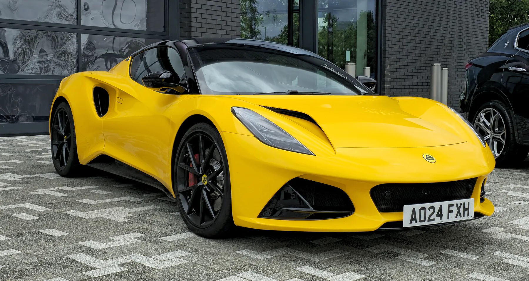 Front three-quarter of a yellow Lotus Emira from the front right, nose to the right, on a gray block-paved forecourt in front of a dark glass-and-brick building. The headlamps are dark. A dark hood duct is visible above the headlamp nearest the camera. A round yellow-and-green Lotus badge sits on the yellow nose above the black lower intake. The plate reads A024 FXH. The roof and mirror are black. A large black intake is behind the door. The wheels are black multi-spoke with red calipers. A dark SUV is at the right edge.
