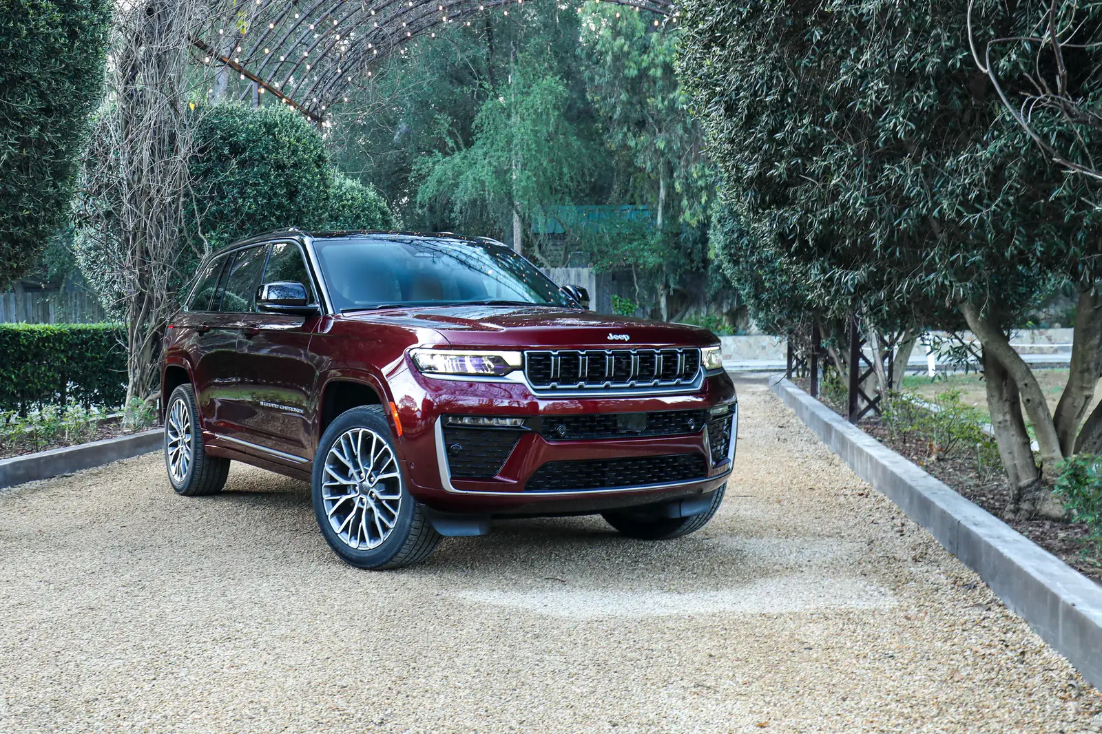 Deep-red 2026 Jeep Grand Cherokee, front three-quarter on gravel under a garden arch, seven-slot grille and slim lamp graphic