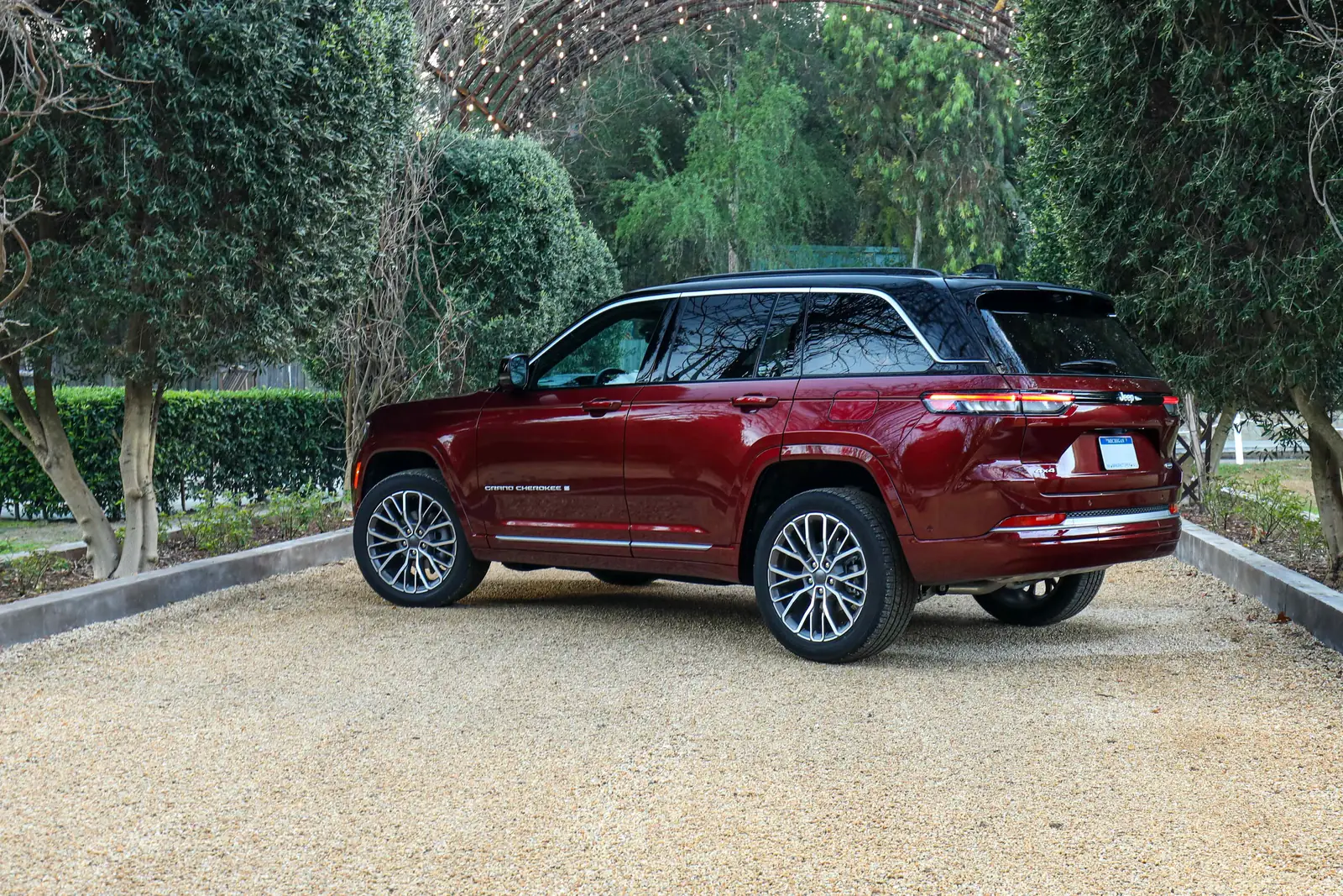 Deep-red 2026 Jeep Grand Cherokee, rear three-quarter on gravel under a garden arch, slim tail lamps