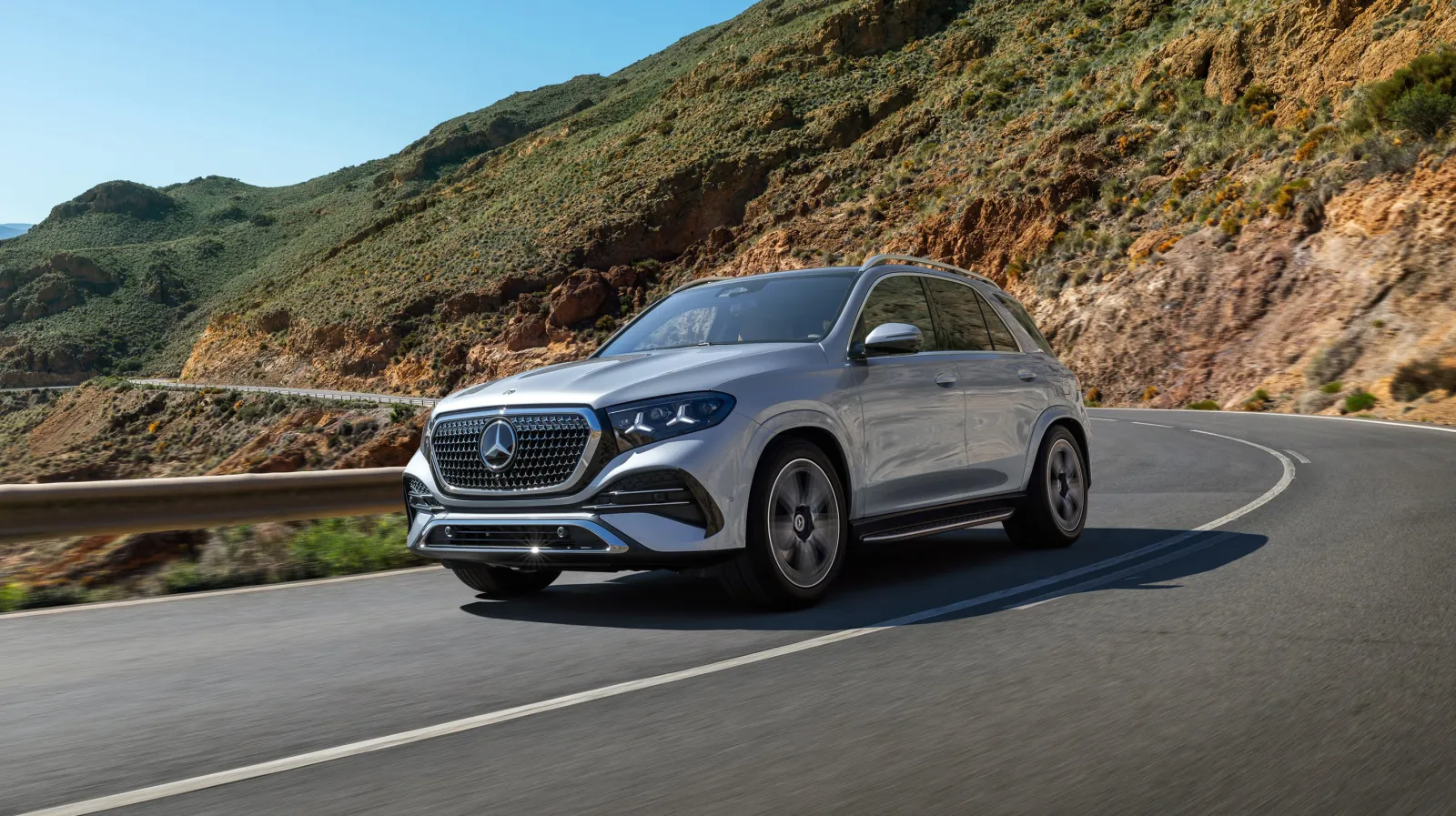 Silver 2027 Mercedes-Benz GLE, front three-quarter on a mountain road with twin-star lamps and a larger grille