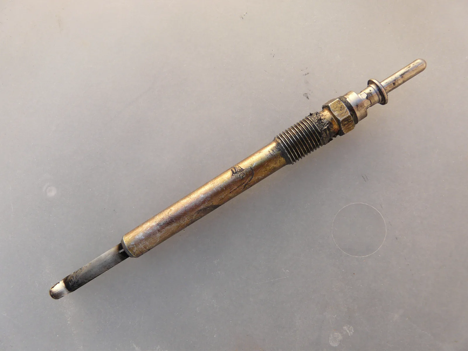 One long glow plug lying diagonally on a plain light-gray surface. The terminal pin with a collar is at the upper right, followed by a brass-colored hex and a short threaded section with dark deposits. A long brass-colored smooth body runs down toward the lower left and ends in a thin gray heater tip, darkened near the body, with a pale end.