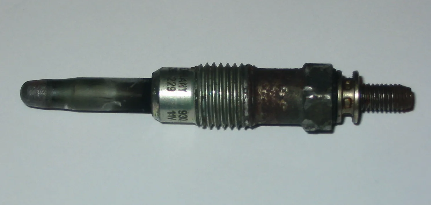 One used glow plug lying horizontally on a pale gray background. At the left is a smooth, unthreaded heater rod with a rounded tip, discolored dark gray. Next is a silver collar with partly visible stamped markings, including characters like 229, 930, and 11V. Then a threaded section, a rust-brown body, and a rusty hex. At the right is a small ring and a threaded terminal stud, with no nut visible.