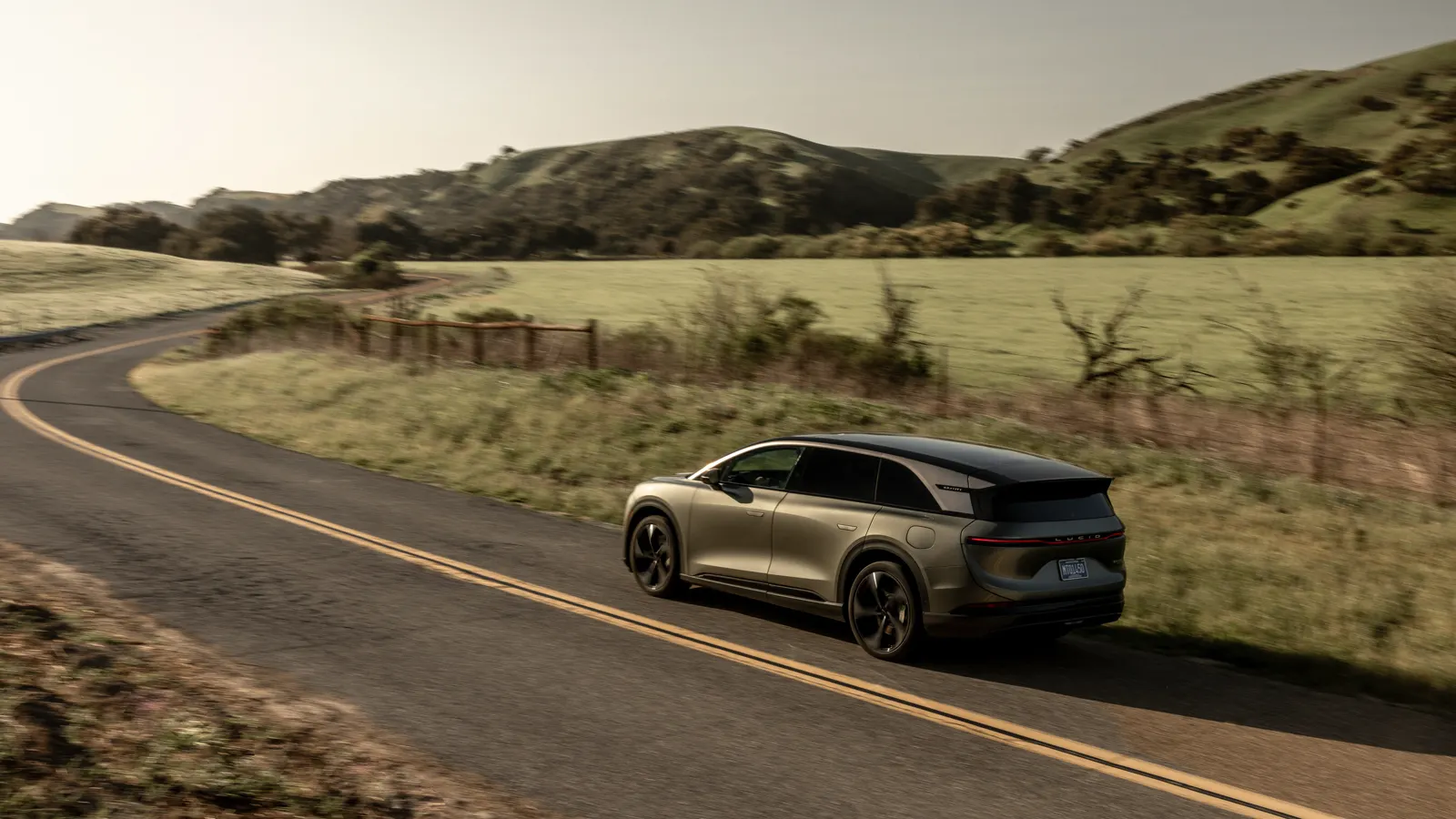 Bronze 2026 Lucid Gravity Grand Touring, high rear three-quarter on a hill road with a glass canopy and slim tail bar