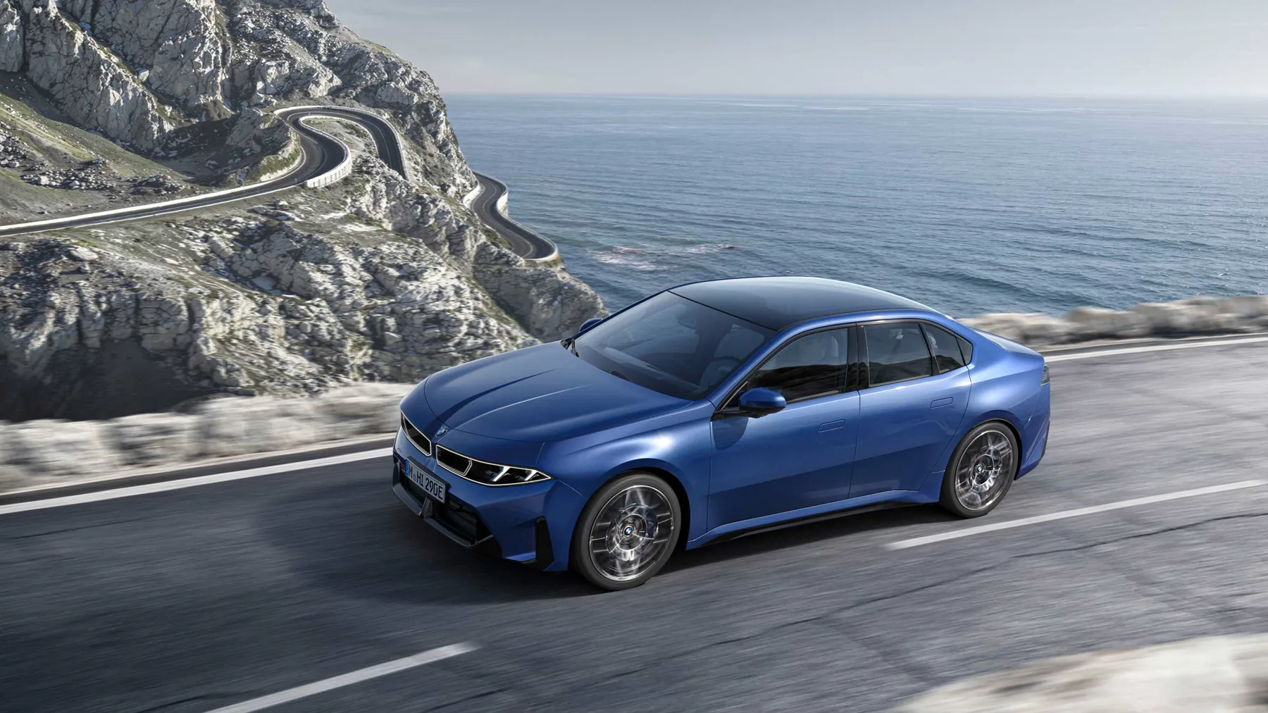 Blue 2026 BMW i3 50 xDrive from above, glass roof and flared arches on a coastal road