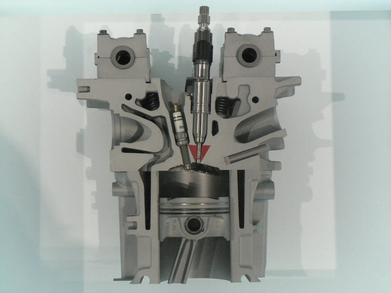 BMW gasoline direct injector seated in a cutaway head, tip aimed at the piston bowl beside a Bosch plug