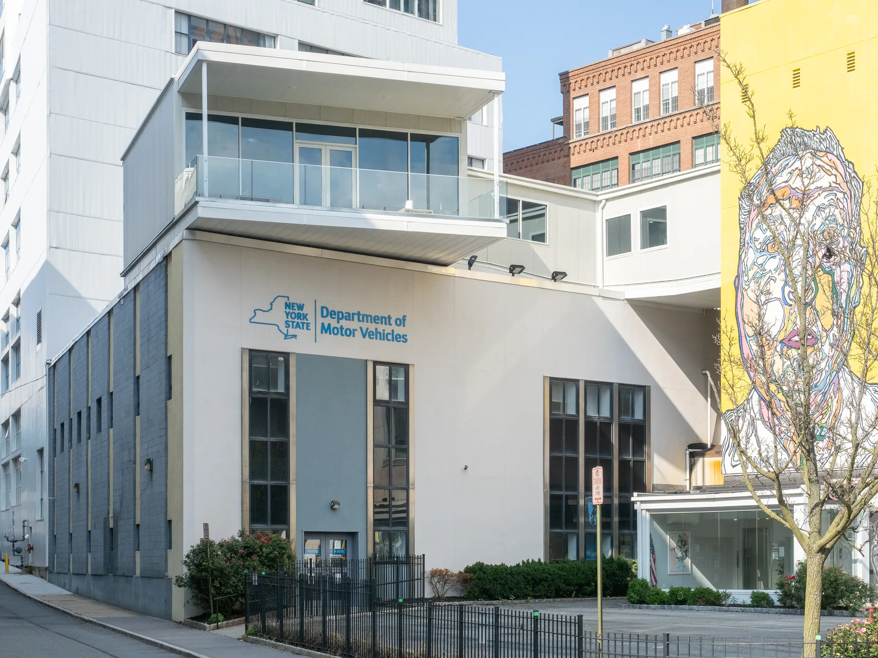 A multi-story white and gray modern building under a clear blue sky. On the facade, a blue New York State outline logo sits beside the words Department of Motor Vehicles. Tall narrow dark windows, a black iron fence and shrubs run along the front, and a sidewalk is at the left. A large painted mural of a face on a yellow wall is at the right, with an older red-brick building behind and a bare young tree in front.