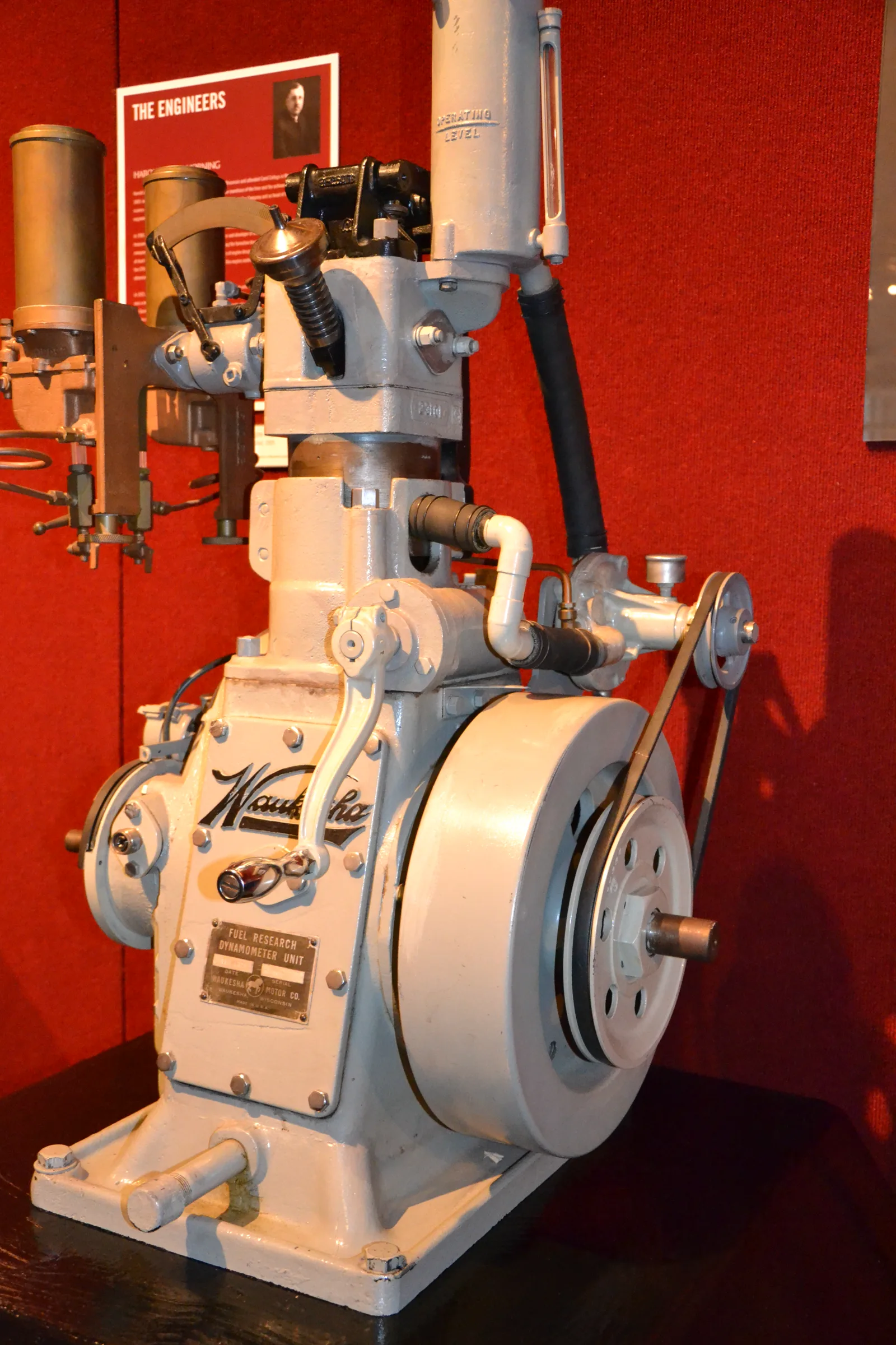 Single-cylinder engine painted pale gray off-white, standing on a dark base against red fabric walls. The script word Waukesha is cast on the crankcase, and a small brass plate below it reads FUEL RESEARCH DYNAMOMETER UNIT. A large solid pale flywheel is at the lower right, with a black belt running up to a small pulley. A black hose and a white pipe are at the right of the cylinder. A tall cylindrical part at the top is marked OPERATING LEVEL and has a sight glass. Brass canisters and linkages are at the upper left, and a red poster headed THE ENGINEERS is on the wall behind