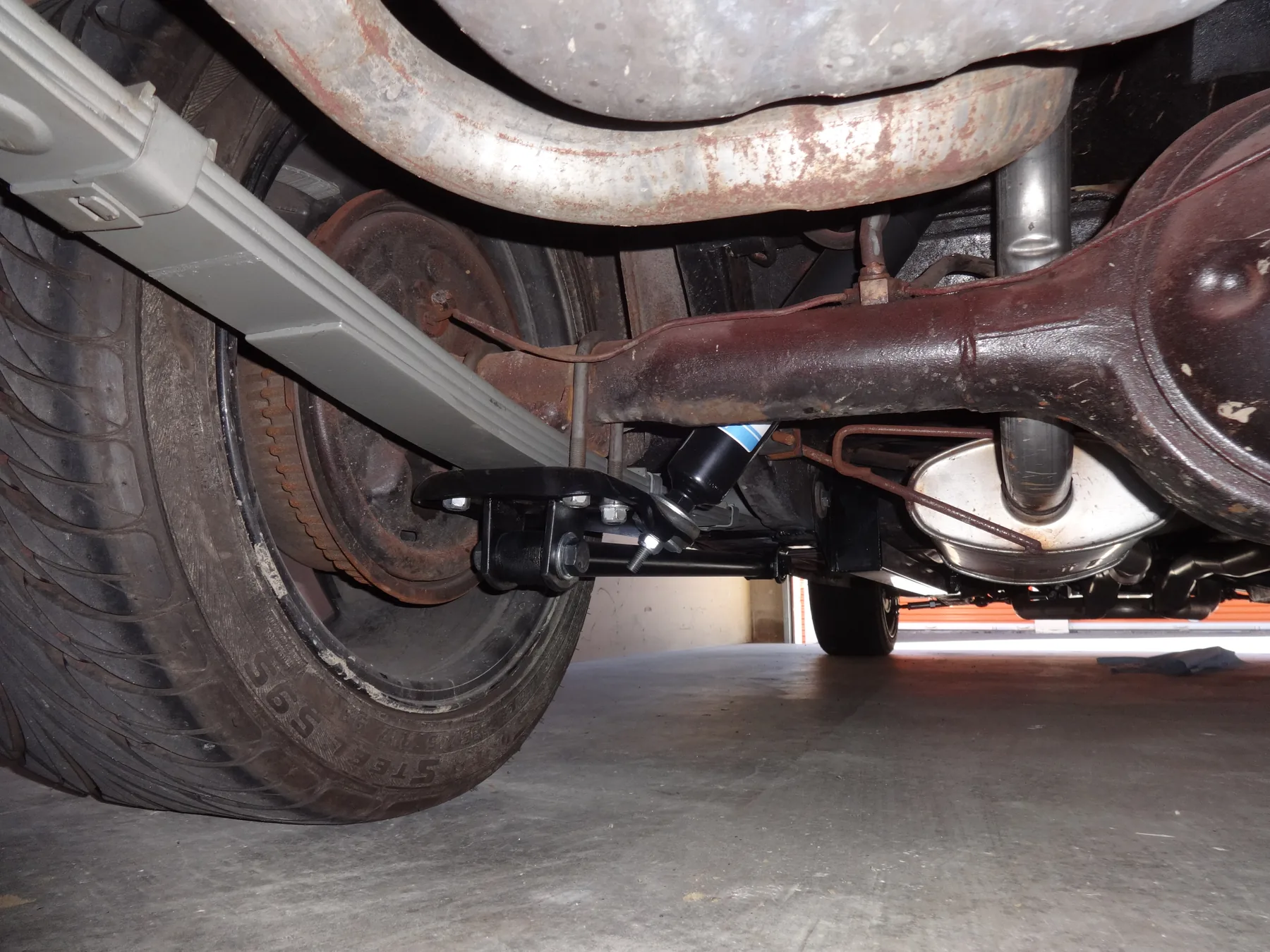Rear underside of a 1965 Ford Mustang with the spring's rear eye in a shackle, the front eye in a frame hanger, U-bolts over the axle, and a shock absorber beside the housing
