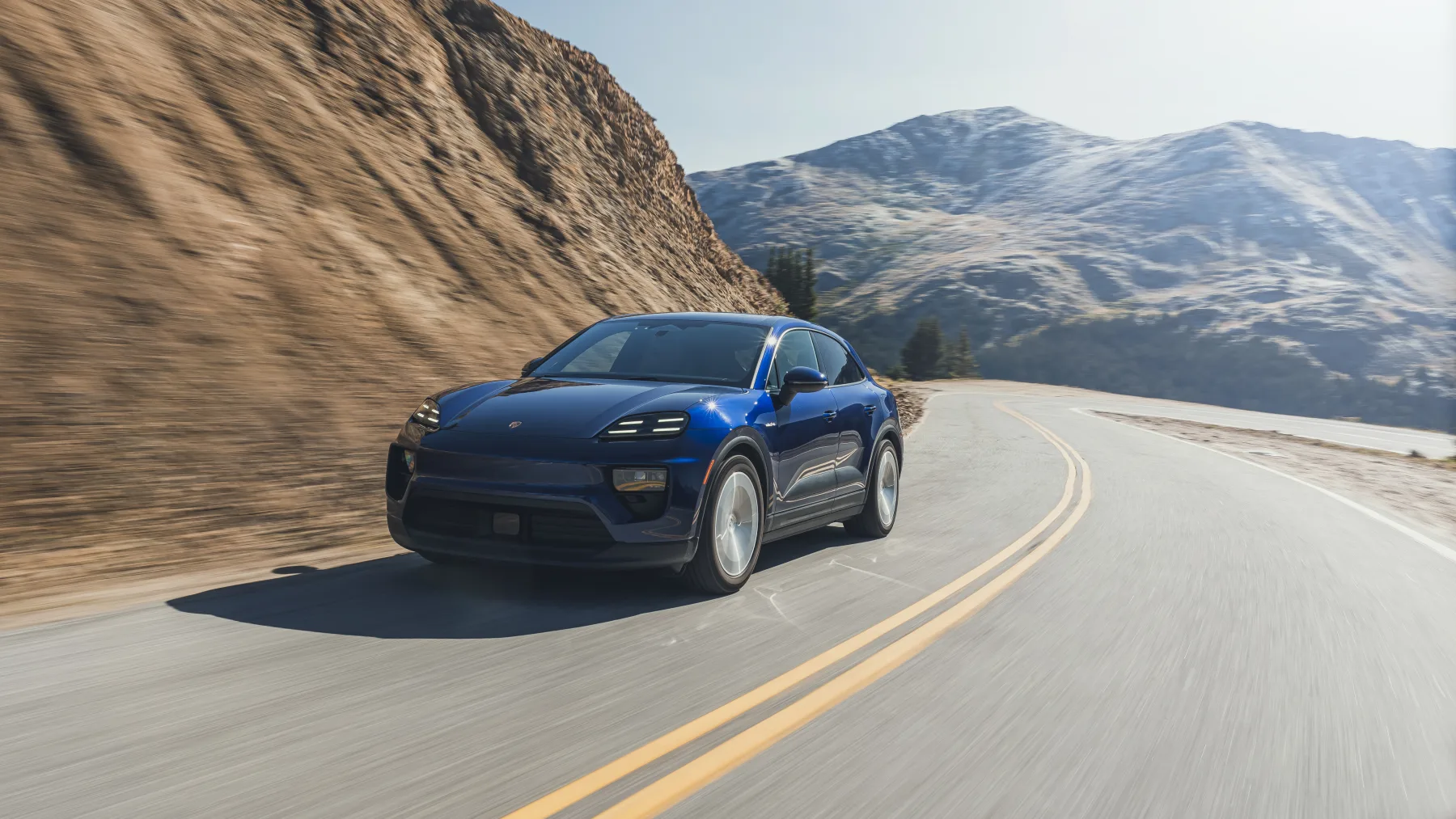 Gentian-blue 2026 Porsche Macan Electric, front three-quarter on a mountain road with four-point lamps lit