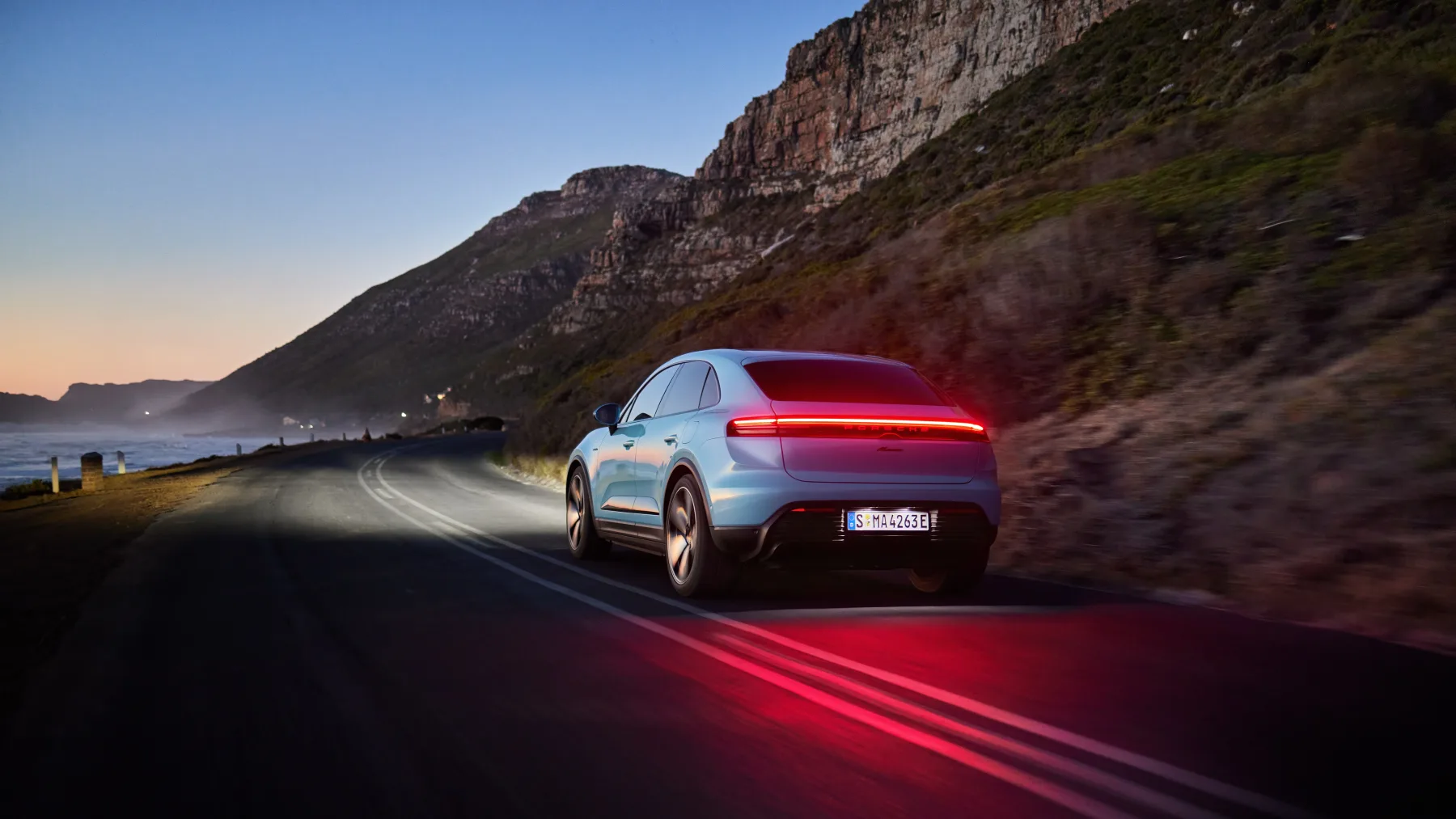 Frozen-blue 2026 Porsche Macan Electric, rear three-quarter with a full-width light bar on a coastal road