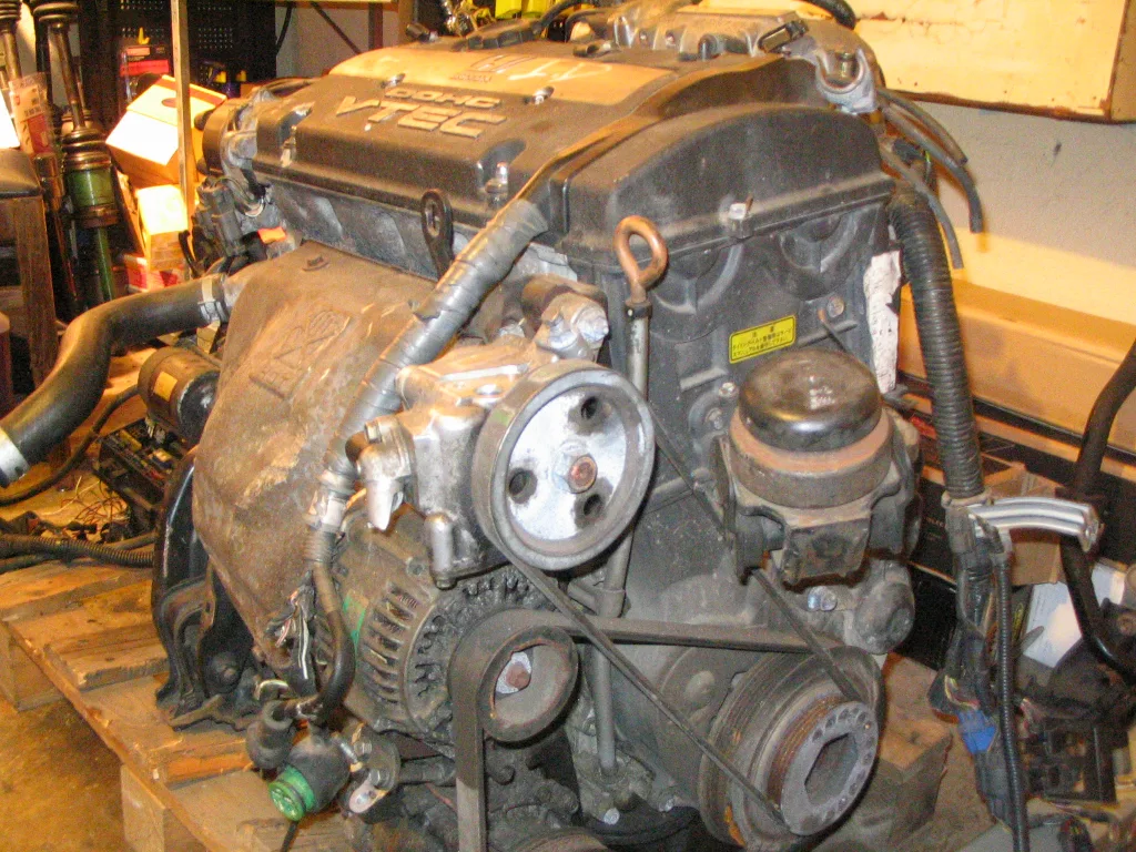 A Honda H22 on a wooden pallet in a cluttered garage. The DOHC VTEC valve cover is on top, a silver power-steering pump pulley is in the middle, an alternator sits below that pulley, a large crank pulley is at the lower right, and a copper-colored dipstick loop rises from the block. Right of center, a black round-topped engine mount sits on a black bracket bolted to the timing-belt side, next to a small yellow warning label.