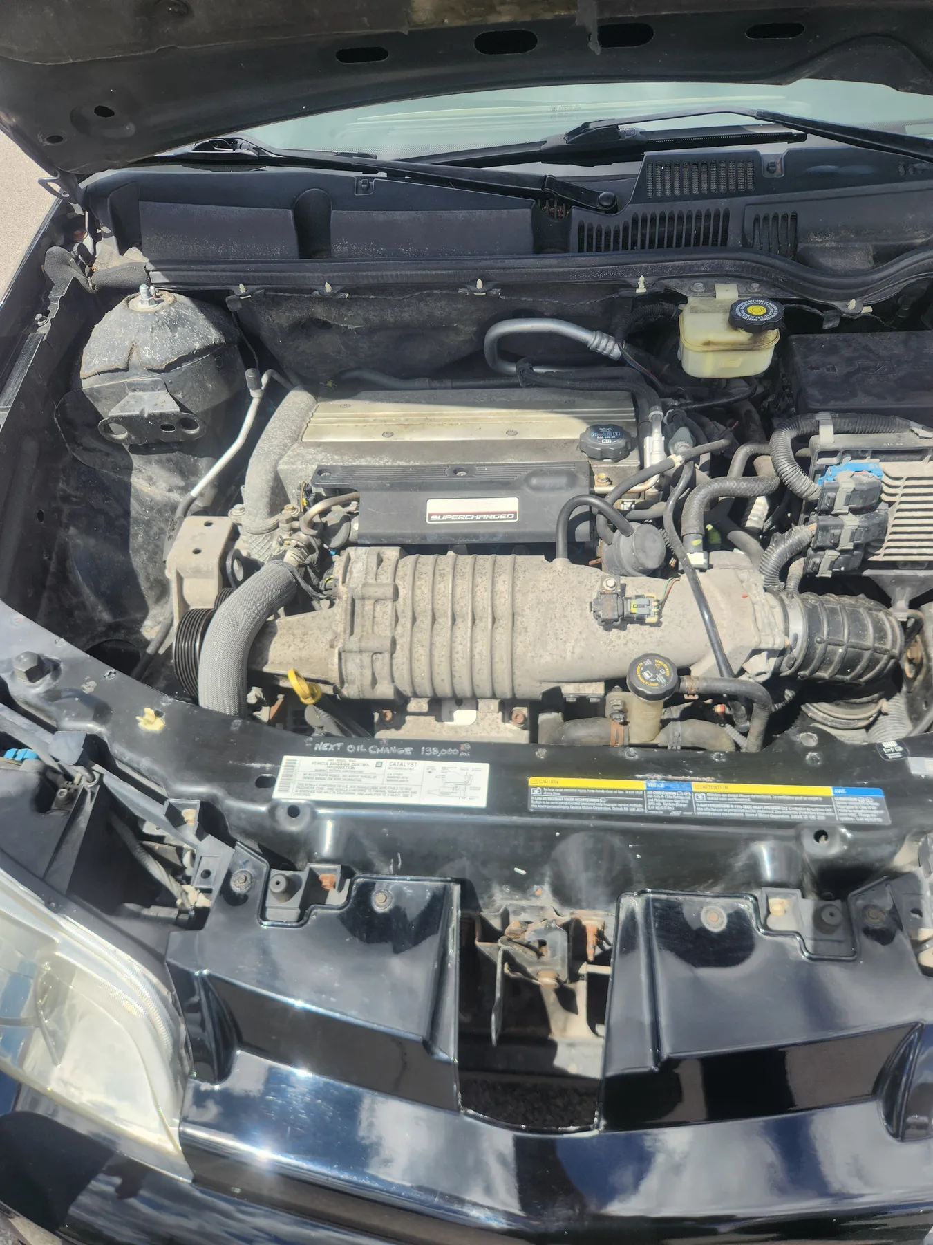 Hood up, looking down into the bay. A gray ribbed supercharger runs across the middle, in front of a black cover badged SUPERCHARGED. A translucent fluid reservoir with a black-and-yellow cap is at the upper right, the cowl and wipers are at the top, and a black radiator support with labels runs across the bottom. The black round mount at the upper left, with a three-hole bracket, is on the frame rail.