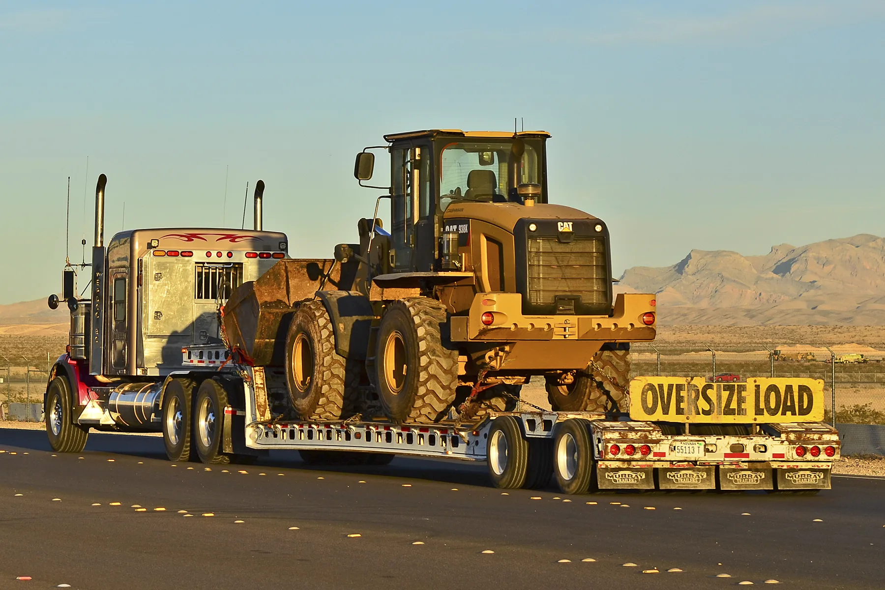 A left-side view of a chrome and silver long-hood semi tractor with a red frame, tall twin exhaust stacks, and a flame stripe on the sleeper. It is pulling a lowboy trailer on a desert highway at low sun, carrying a yellow CAT wheel loader with its bucket toward the cab. A yellow sign with black letters reading OVERSIZE LOAD is mounted at the rear of the trailer, below the back of the loader and above the red tail lamps. Bare mountains are behind.