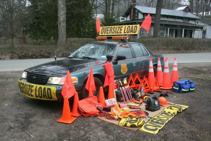 A dark green and black Ford Crown Victoria-style sedan from the front left, parked on dirt by a road. It has a yellow OVERSIZE LOAD sign on the roof with two orange flags and an amber light bar, an OVERSIZE LOAD banner across the front bumper, and a gold badge-shaped decal on the front door. Escort gear is laid out on the ground beside it: orange traffic cones, orange flags, red reflective triangles, folded yellow OVERSIZE LOAD banners, a first-aid kit, and a blue bag. A house with a green roof and bare trees are behind.