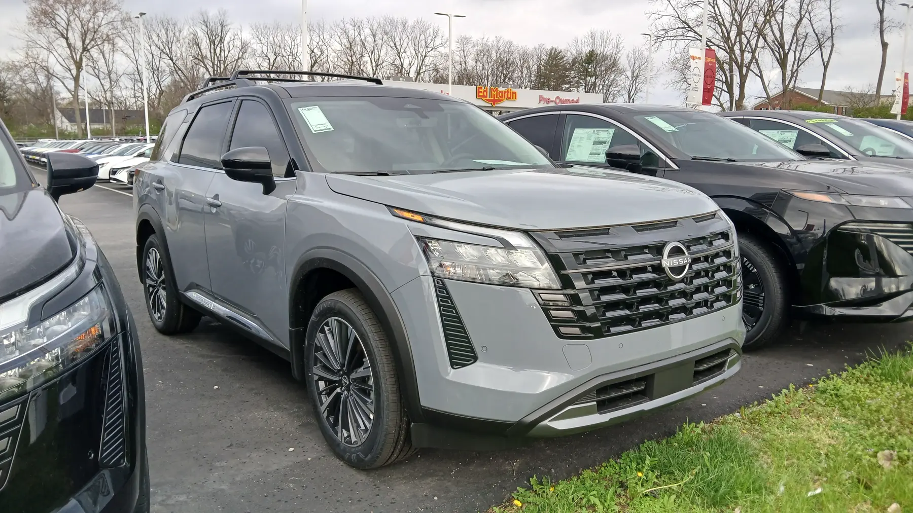 Front three-quarter of a gray Nissan Pathfinder in a dealer lot, nose to the right. The hood is smooth. The grille is dark horizontal slats with bright bars, and the roundel is bright with NISSAN across the bar. The headlamp is a clear main lens with a thin element above it and an amber segment at the outer corner. The lower bumper is body color, with dark openings. A bright strip along the lower door reads PATHFINDER. Black roof rails run above the glass. A window sticker is on the windshield. The sign behind reads Ed Martin and Pre-Owned. The wheels are multi-spoke with bright faces. Other vehicles fill the lot, a black SUV is at the right, and grass is in the foreground.