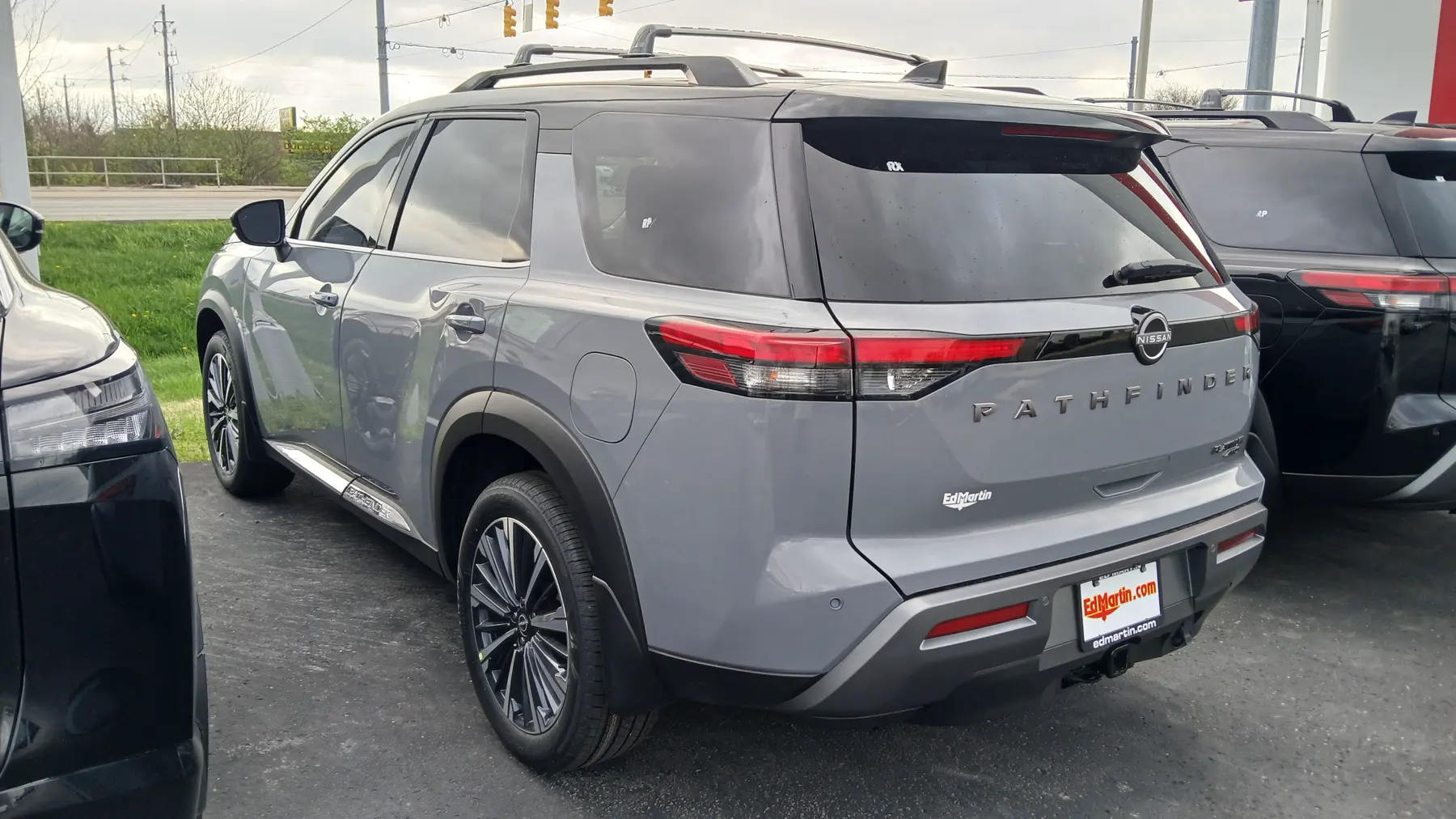 Rear three-quarter of a gray Nissan Pathfinder in a dealer lot, tail toward the right and the flank to the left. Spaced metal letters spelling PATHFINDER run across the liftgate, with a Nissan roundel above them and a small PLATINUM badge at the lower right of the gate. A red lamp crosses the gate, with a clear section in the lens. A white Ed Martin decal is on the liftgate, at the left below the lamp. The rear side glass and the liftgate glass are dark, and a fuel door is on the quarter. A bright strip runs along the lower doors. Black roof rails run above the glass. The rear bumper is body color with a dark lower section, a red reflector, and a hitch. The plate frame reads EdMartin.com and edmartin.com. The wheels are multi-spoke with bright faces. A black SUV is parked at the right, and grass and a road are behind.
