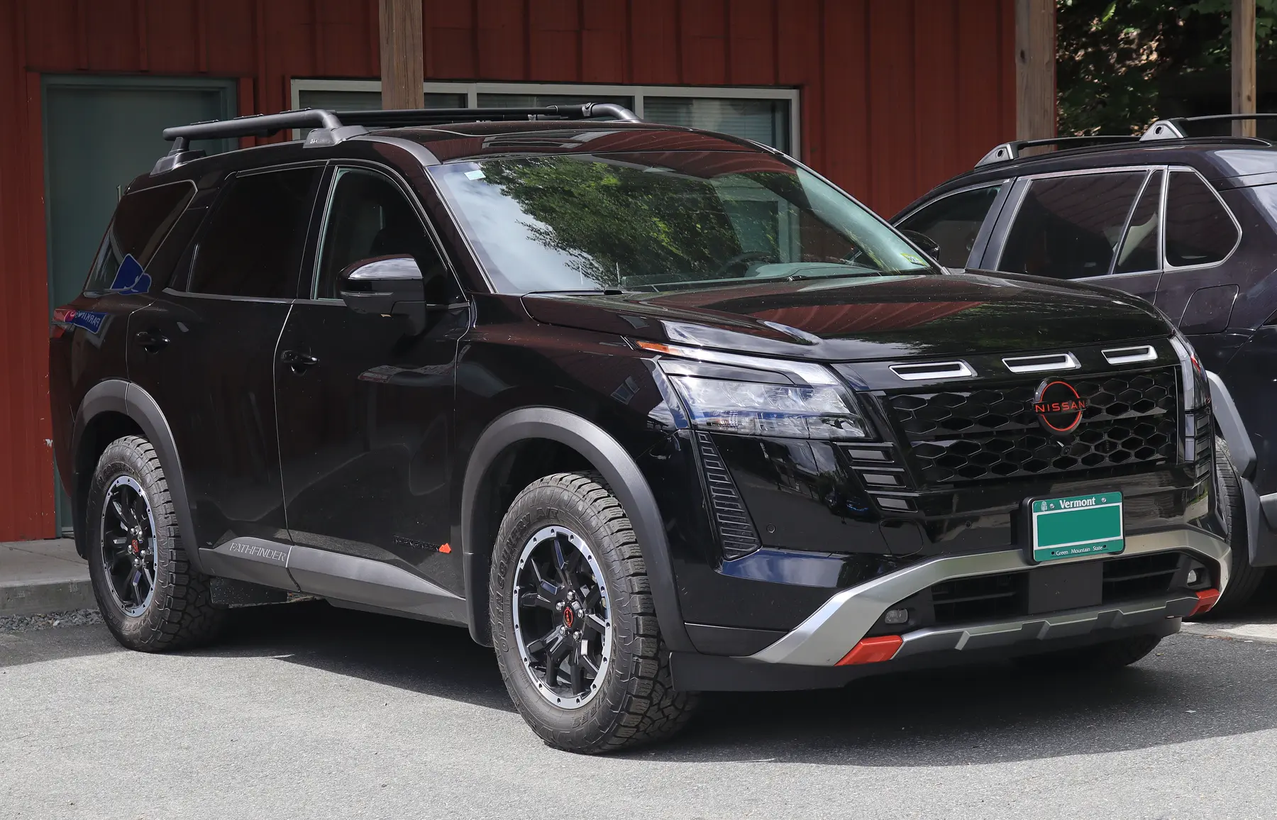 Front three-quarter of a black Nissan Pathfinder on asphalt, nose to the right, in front of a red building with a pale door and windows. Three bright rectangular vents sit on the hood. The grille is a black honeycomb mesh in a bright surround, and the roundel has a red ring with NISSAN across the bar. The headlamp is a clear main lens with a thin element above it and an amber segment at the outer corner. The lower bumper is silver, with red pieces at the outer corners. A green plate reads Vermont at the top and Green Mountain State along the bottom. Black roof rails run above the glass, with a crossbar. The front fender reads ROCK CREEK in dark letters beside a small red mark, and the lower cladding reads PATHFINDER in light letters. The wheel is black with a bright ring of fasteners and a red center cap, and the tire tread is blocky. A dark SUV is parked at the right.