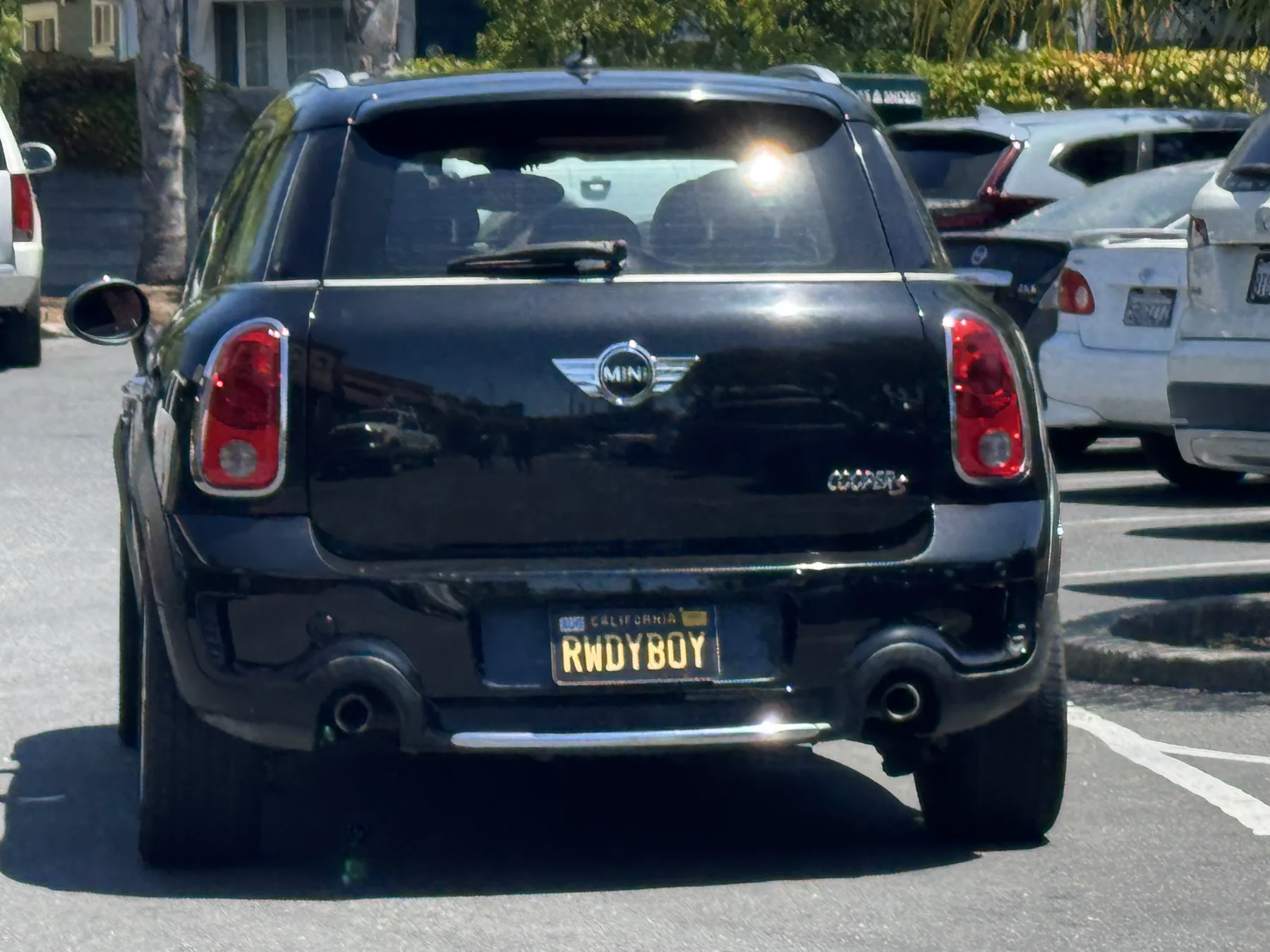 Straight-on rear of a black MINI Cooper Countryman in a sunny parking lot. A COOPER S badge and the MINI wing badge are on the hatch. Red-and-white vertical taillamps sit at the upper corners, with twin exhaust tips and a chrome strip on the lower bumper. The plate is a black California legacy plate with gold characters reading RWDYBOY and CALIFORNIA in small gold letters at the top, mounted low in a recess in the bumper. A white SUV is at the left edge, white sedans are parked at the right, and hedges and a building are behind.