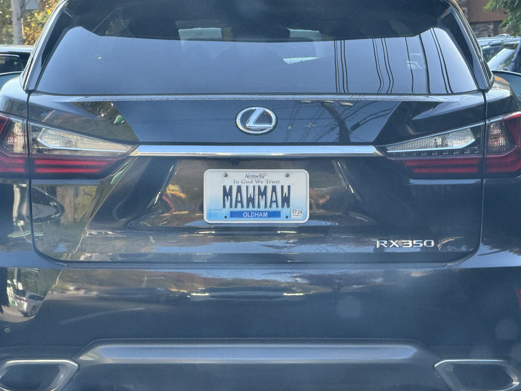 Straight-on rear of a dark Lexus RX 350. The Lexus badge is above the plate and an RX350 badge is at the lower right. Wide wraparound taillamps and a chrome strip run across the hatch, with two exhaust outlets at the bottom. The white Kentucky plate reads MAWMAW in dark letters, with Kentucky script and In God We Trust across the top, a blue band reading OLDHAM at the bottom, and a 07/24 sticker at the lower right.