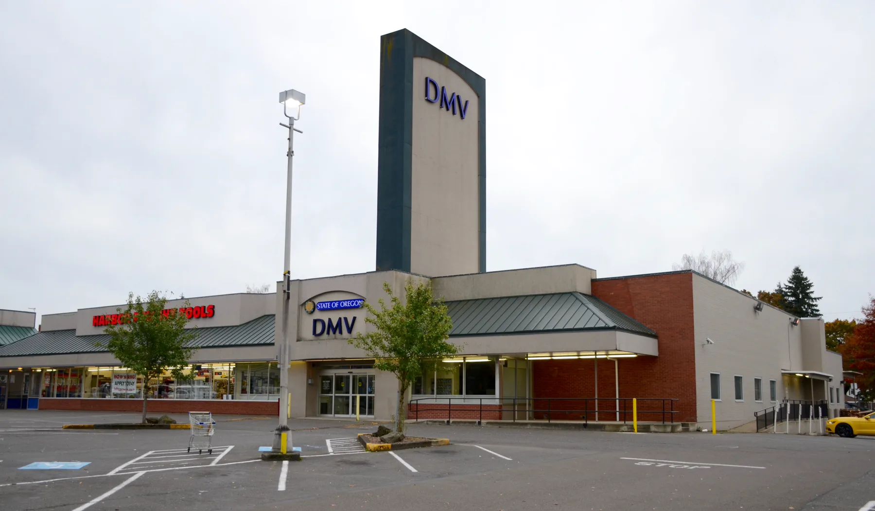 An Oregon DMV office in a strip-mall lot under an overcast gray sky. A tall gray-and-dark-teal pylon reads DMV in blue. Over the entrance a sign reads STATE OF OREGON above DMV. Green standing-seam metal roof sections, a red-brick wall section at the right, and a Harbor Freight Tools sign at the left. A light pole and a shopping cart are in the foreground, a yellow car is at the right edge, and autumn trees are behind.