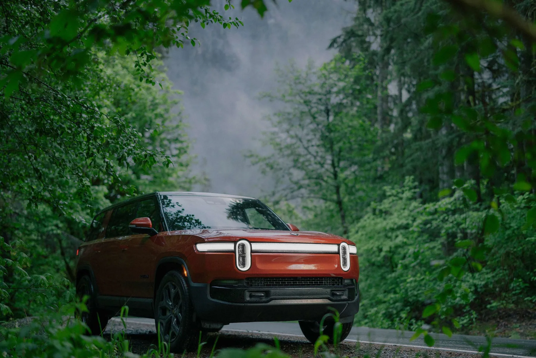 Canyon-orange 2026 Rivian R1S, low front three-quarter in a forest with vertical stadium lamps lit