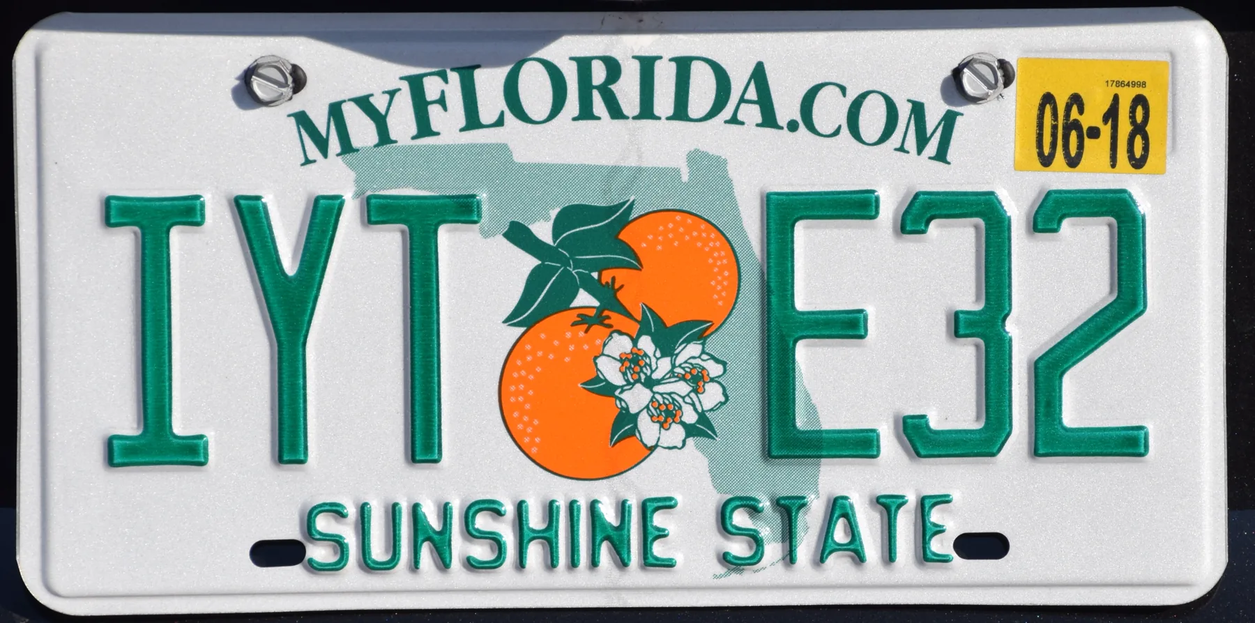 A white Florida license plate photographed straight on, with green characters reading IYT E32. A pale green outline of the state sits behind two oranges, green leaves, and white blossoms. MYFLORIDA.COM is printed in green along the top and SUNSHINE STATE along the bottom. Two metal fasteners fill the top mounting holes, and two empty slots sit along the bottom edge. A yellow sticker in the upper right reads 06-18 in large black digits, with 17864998 in smaller black digits above them. The background is dark.