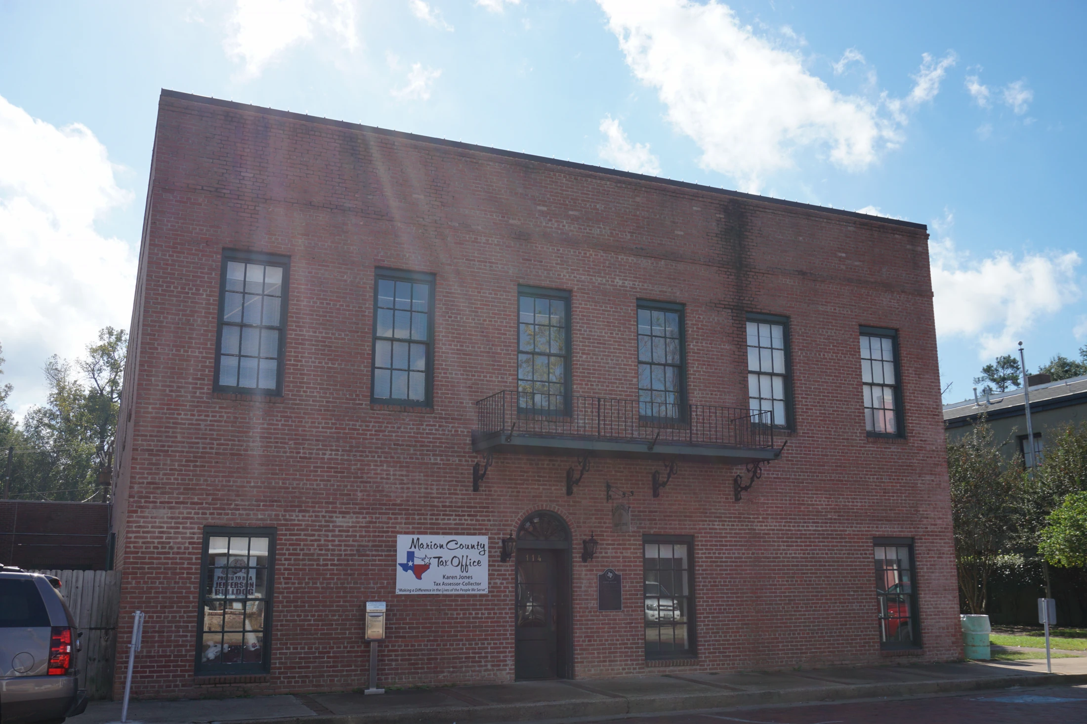 A two-story red brick building under a blue sky with white clouds, seen from the street, with sun glare on the upper left brick. A white sign left of a dark door reads Marion County Tax Office beside a Texas outline and a star, then Karen Jones, Tax Assessor-Collector, and the line Making a Difference in the Lives of the People We Serve. The door is numbered 114, with a fanlight and a black lantern on each side. A brass mailbox stands on a post to the left of the sign. A dark plaque with a Texas outline is on the wall to the right of the door. An iron balcony crosses the second floor. The rear corner of a gray SUV, its taillight showing, is at the left edge, and a green barrel is at the right.