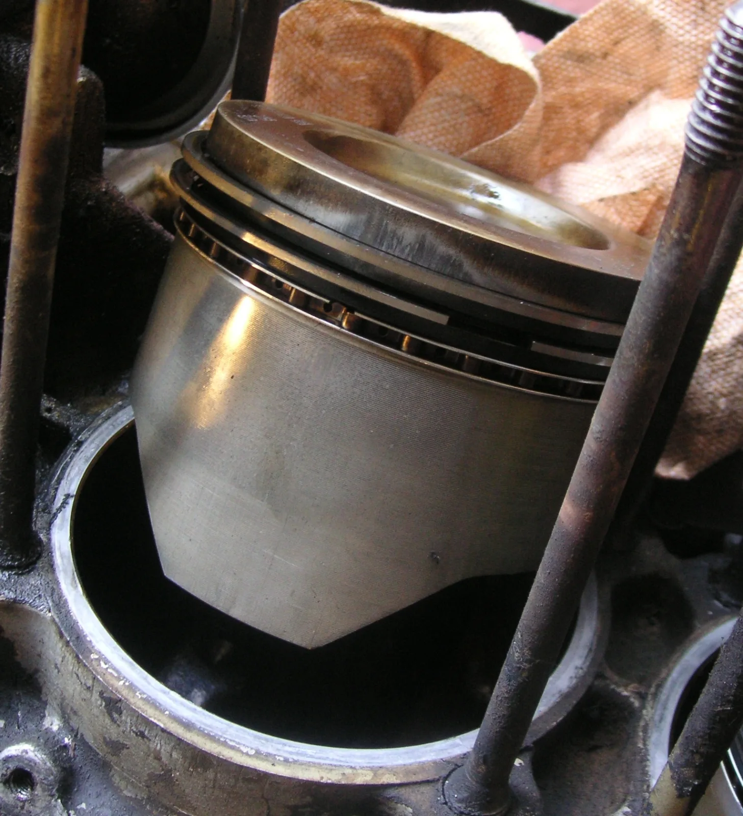 A silver piston tilted and partly lowered into an open cylinder bore in a dirty engine block. Rusty long studs stand on both sides, and a tan shop rag is behind the piston. The upper grooves hold two thin compression rings and, below them, a slotted oil-control ring.