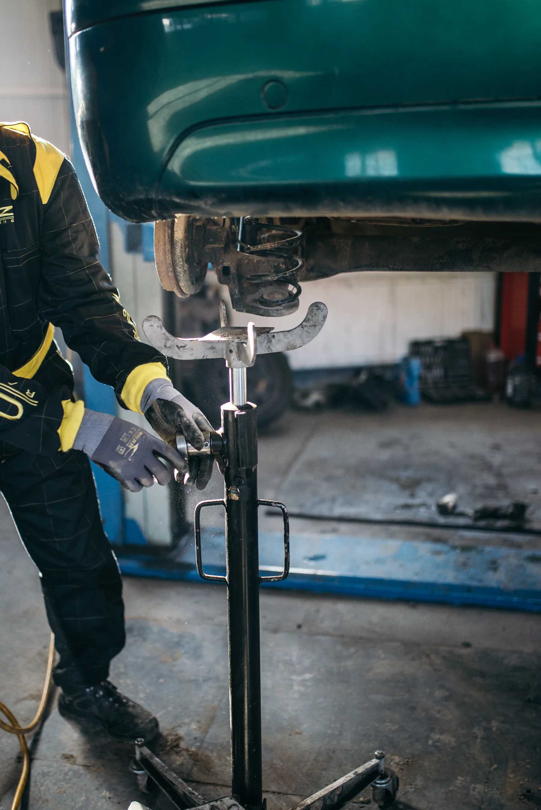 Inside a shop, a person in a black coverall with yellow shoulders and yellow cuffs, gray gloves, and black shoes stands at the left with both hands on a fitting of a black wheeled stand. The stand's silver saddle, two curved arms, sits under a raised dark green car. A coil spring and a dirty brake drum show in the underbody. The floor is stained concrete, a yellow hose lies at the lower left, and a blue lift rail runs along the floor behind.