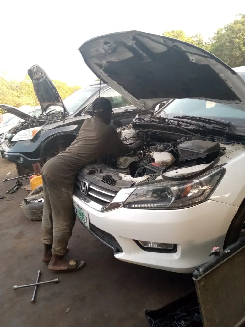 Outdoors in daylight, a person in an olive-brown coverall, a black cap, and sandals leans into the open engine bay of a white car. A Honda emblem is centered on the chrome grille. A dark vehicle behind it also has its hood up. A cross-shaped lug wrench lies on the dirt at the lower left, and the edge of a dark radiator core is at the lower right. Trees fill the background.