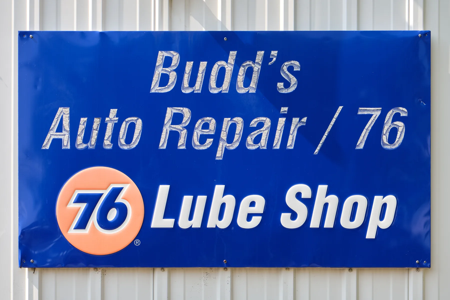 A blue rectangular sign fastened to a white vertically ribbed metal wall. Worn silver-white letters read Budd's on the top line, Auto Repair / 76 on the middle line, and Lube Shop on the bottom line. An orange circle at the lower left contains a blue 76, with a small registered-trademark mark beside the circle.